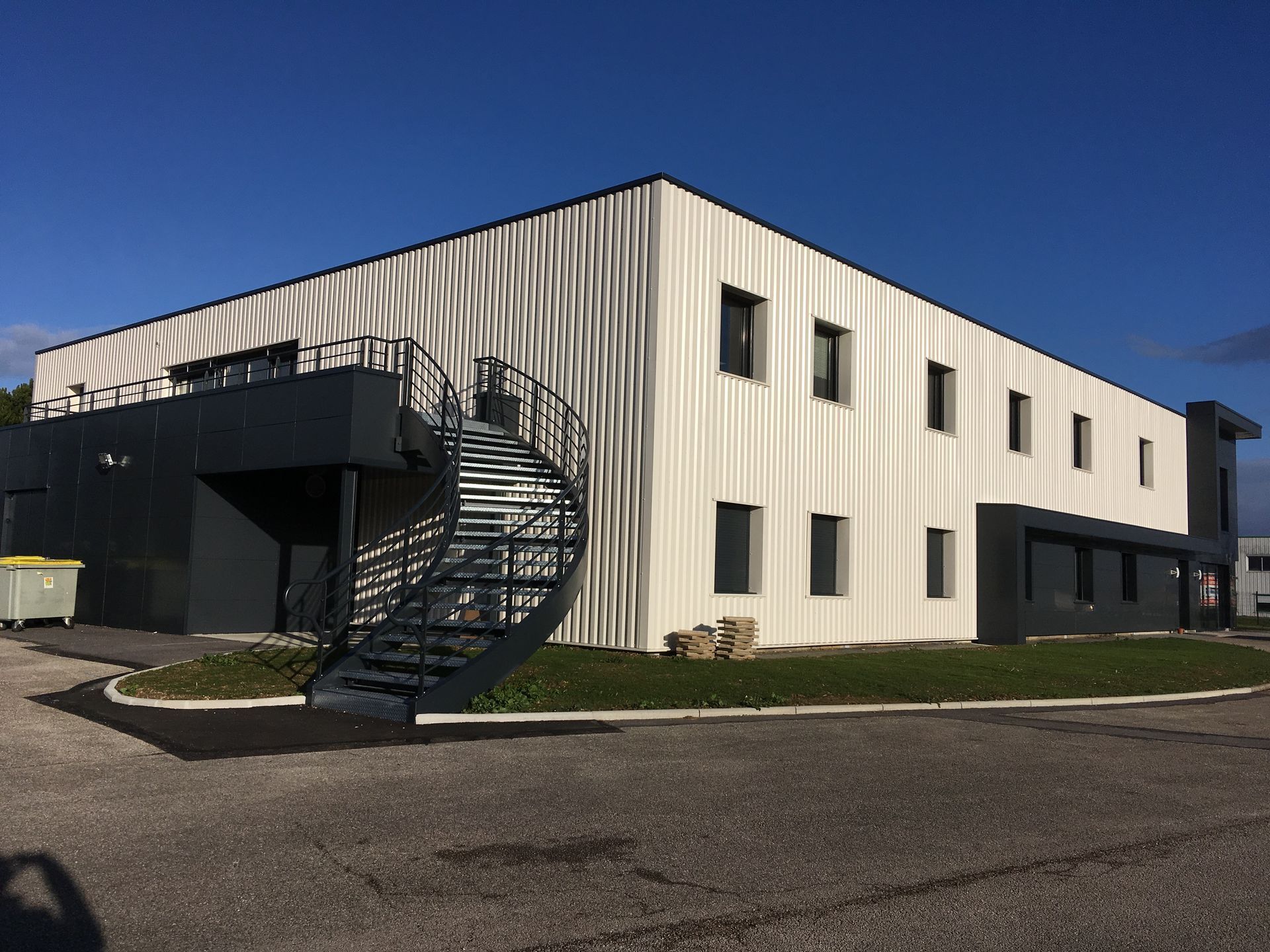 Bâtiment industriel moderne avec une façade en métal beige et des fenêtres. Le bâtiment est entouré de verdure et le ciel est clair et ensoleillé.