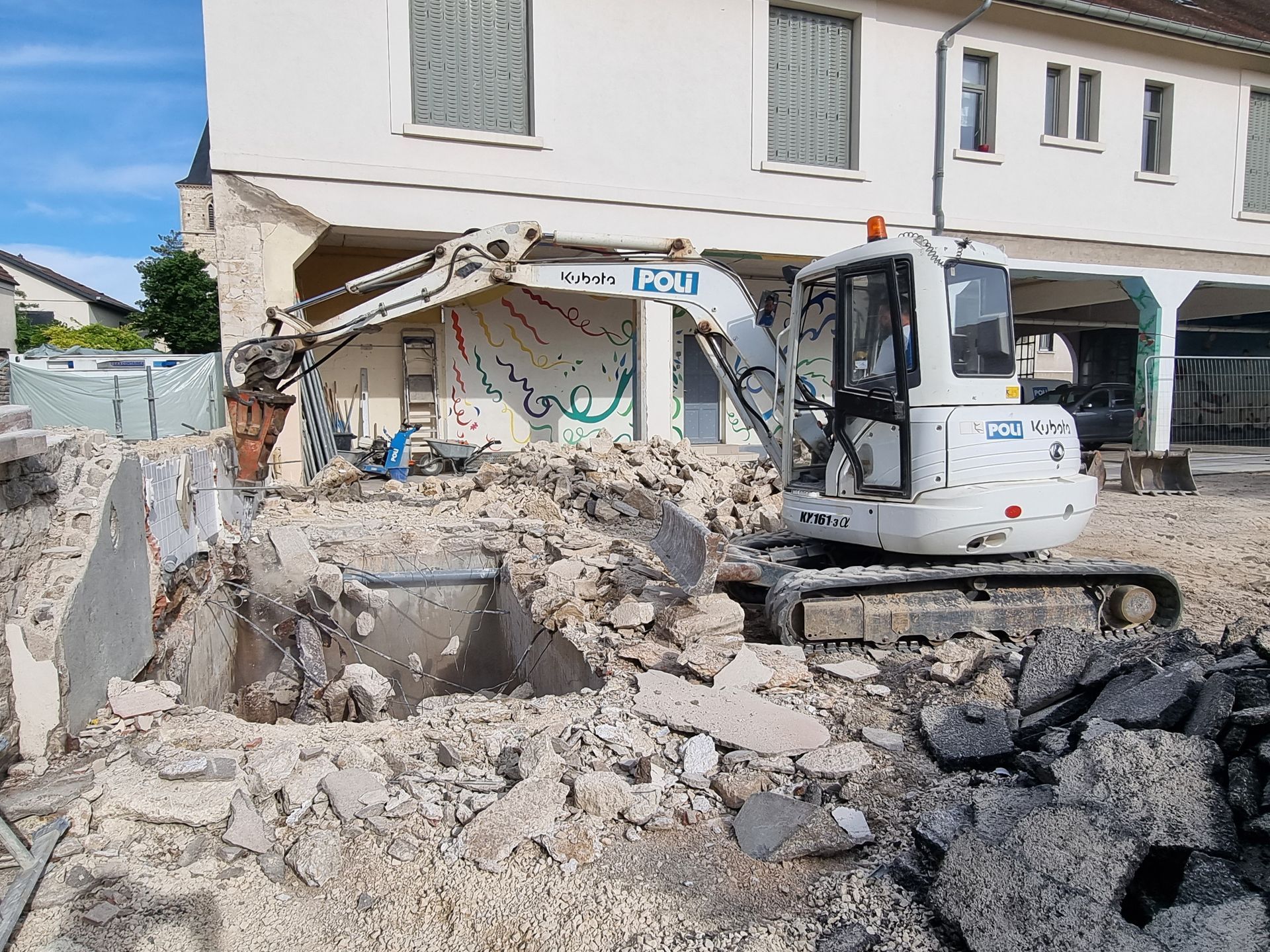 Chantier de démolition avec une pelleteuse en train de travailler sur des débris de construction. Des structures en béton et en métal sont visibles parmi les débris.