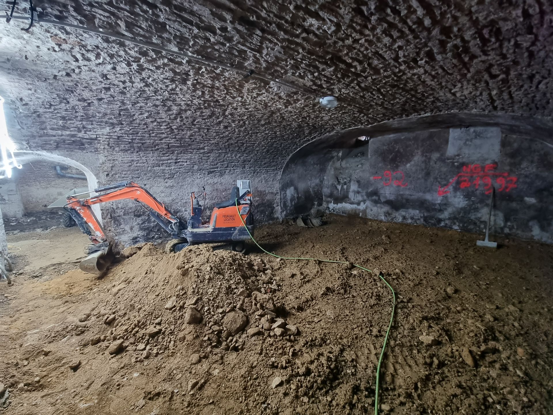 Chantier de construction souterrain avec un engin de chantier en train de creuser. Des structures en métal et des tuyaux sont visibles.