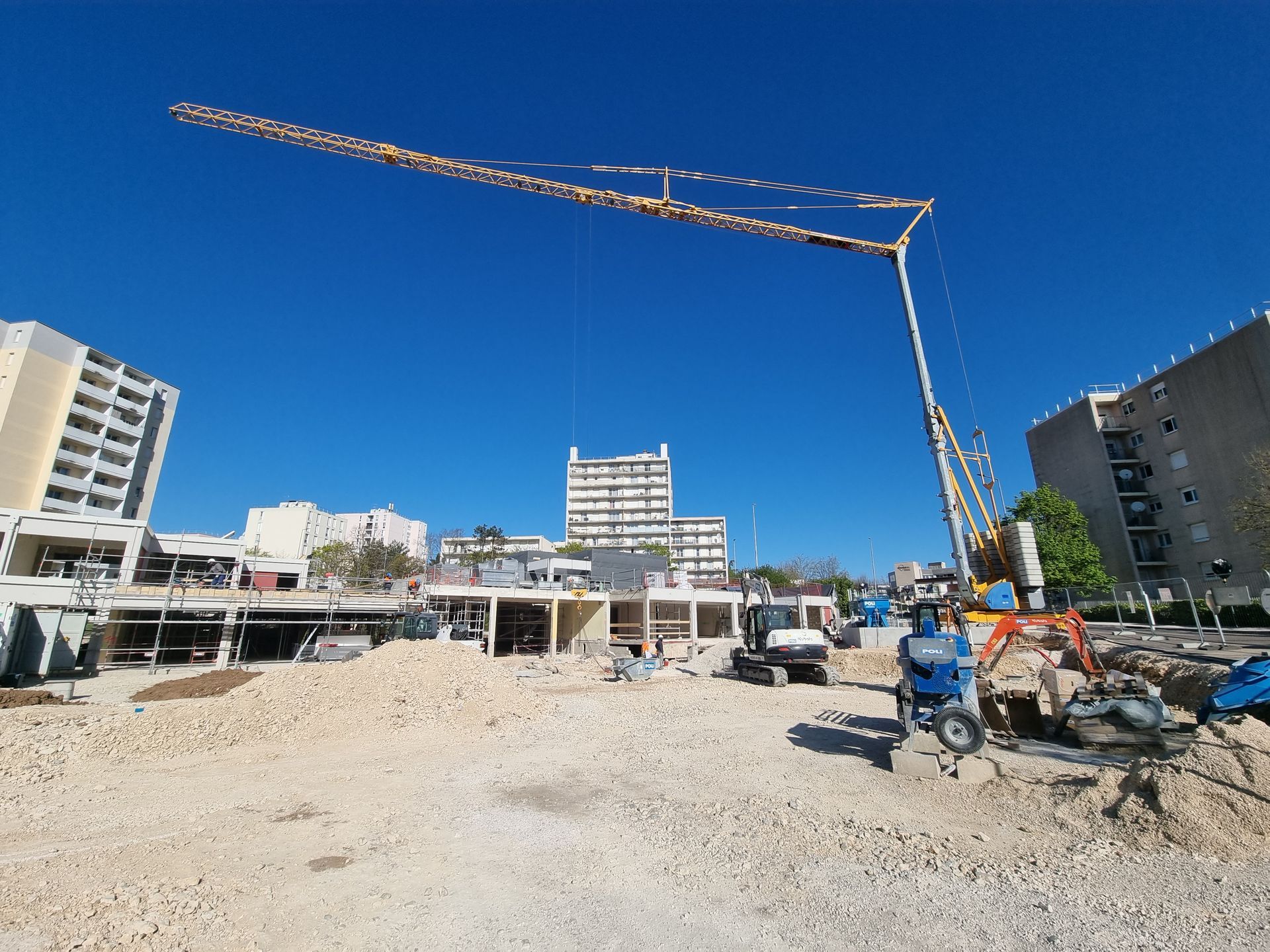 Grue de construction sur un chantier avec des bâtiments en arrière-plan. Le ciel est clair et ensoleillé.