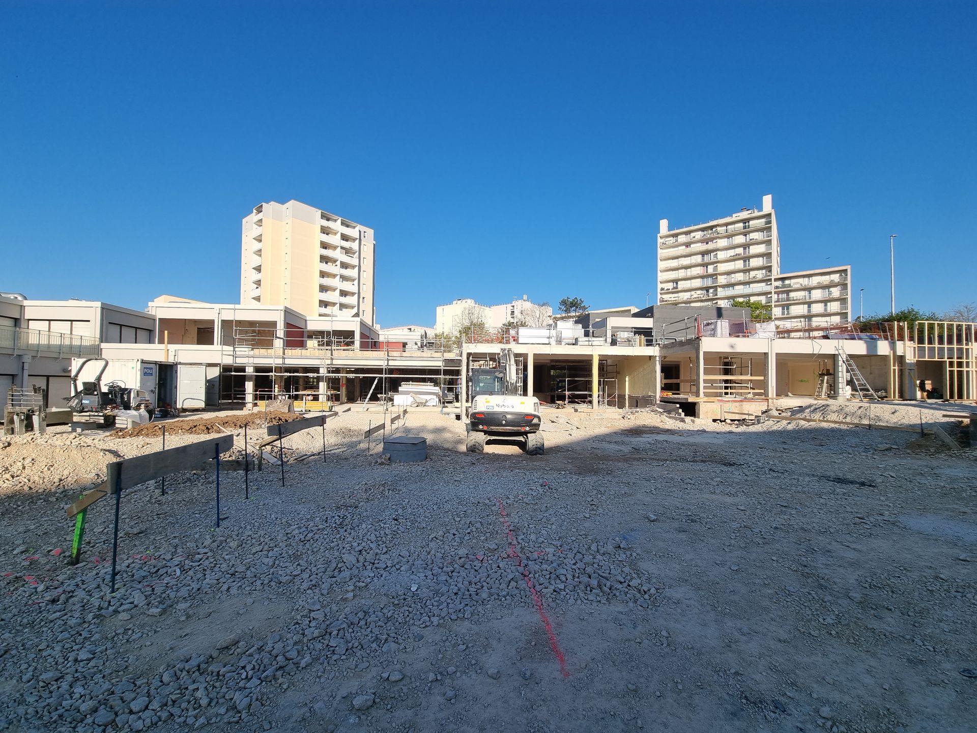 Chantier de construction avec des matériaux de construction éparpillés sur le sol. Des bâtiments en arrière-plan sont partiellement construits.