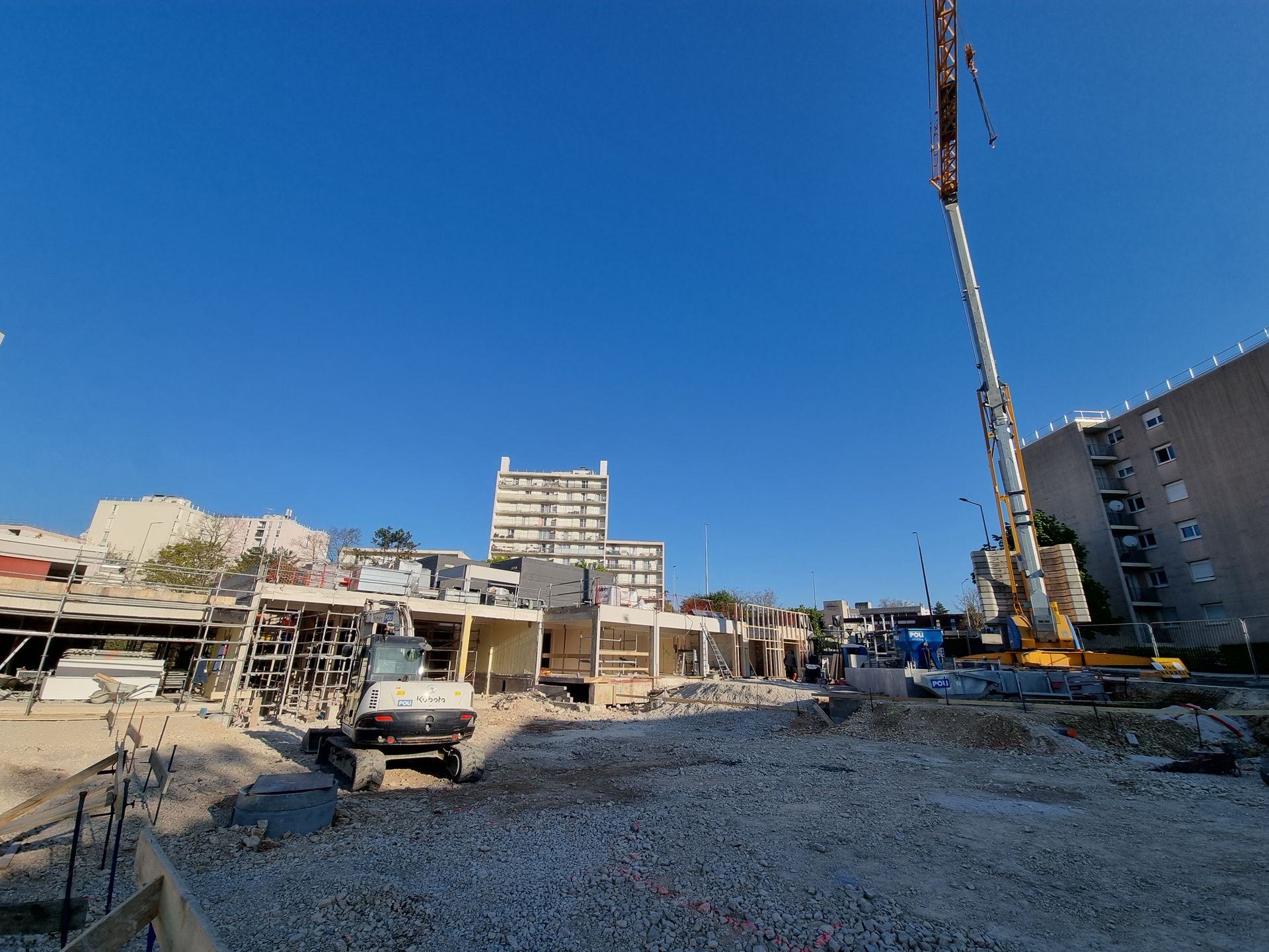Grue de construction sur un chantier avec des bâtiments en arrière-plan. Le ciel est clair et ensoleillé.