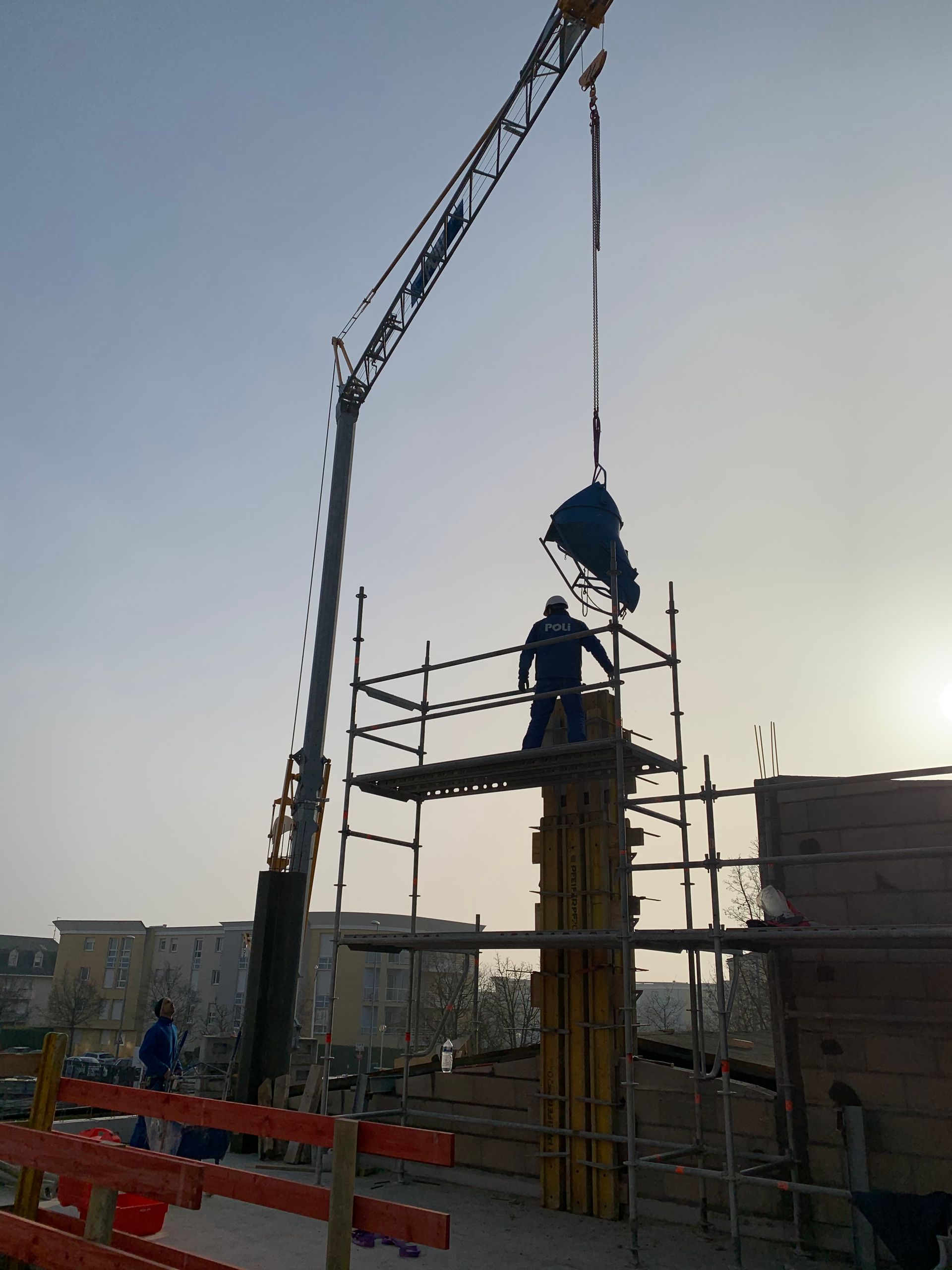 Grue de construction soulevant des matériaux sur un chantier. Un travailleur est visible en haut de la grue, et le ciel est clair avec un éclairage de jour.