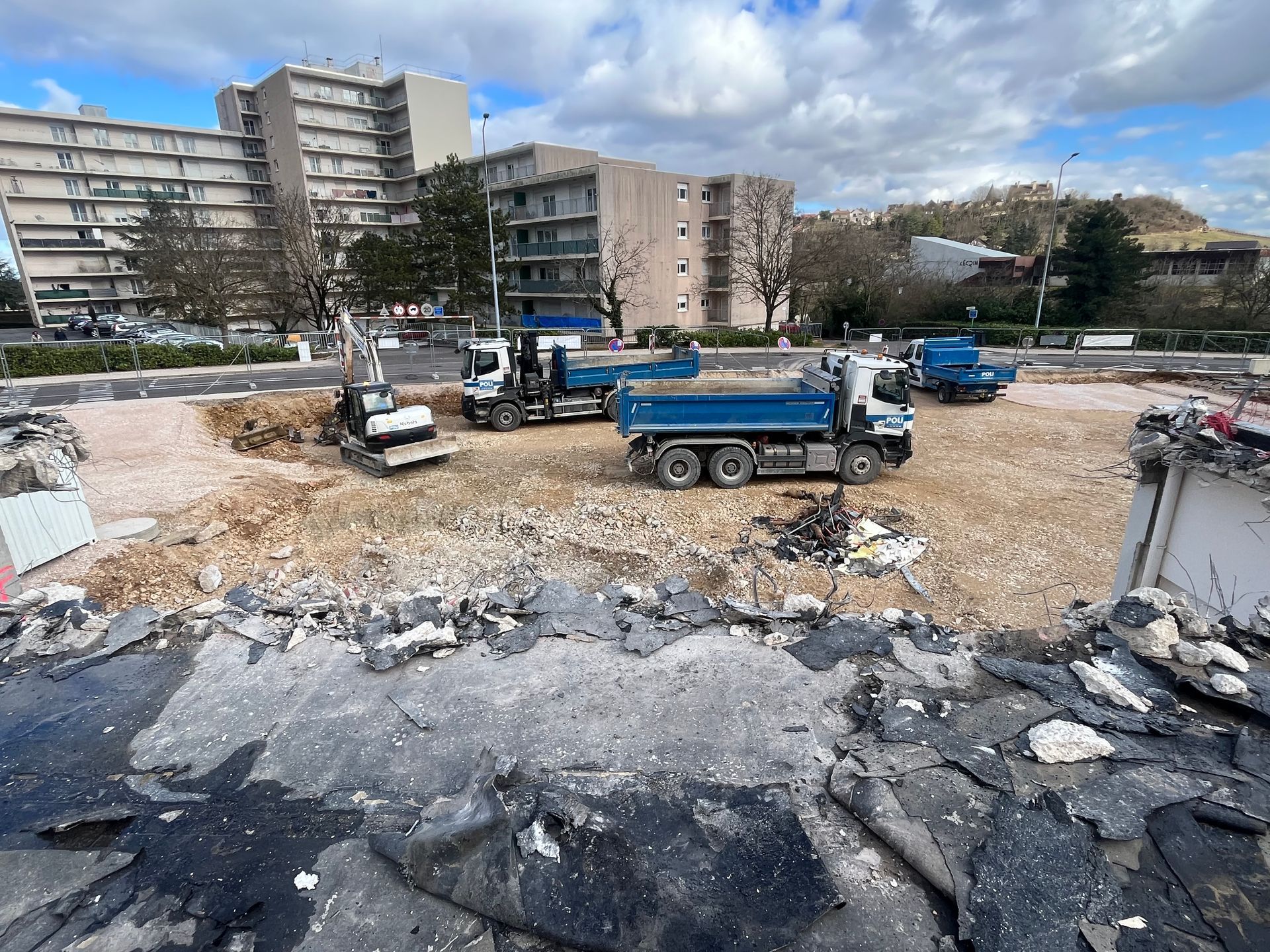 Chantier de démolition avec des débris de construction et un engin de chantier. Des bâtiments en arrière-plan sont partiellement démolis.
