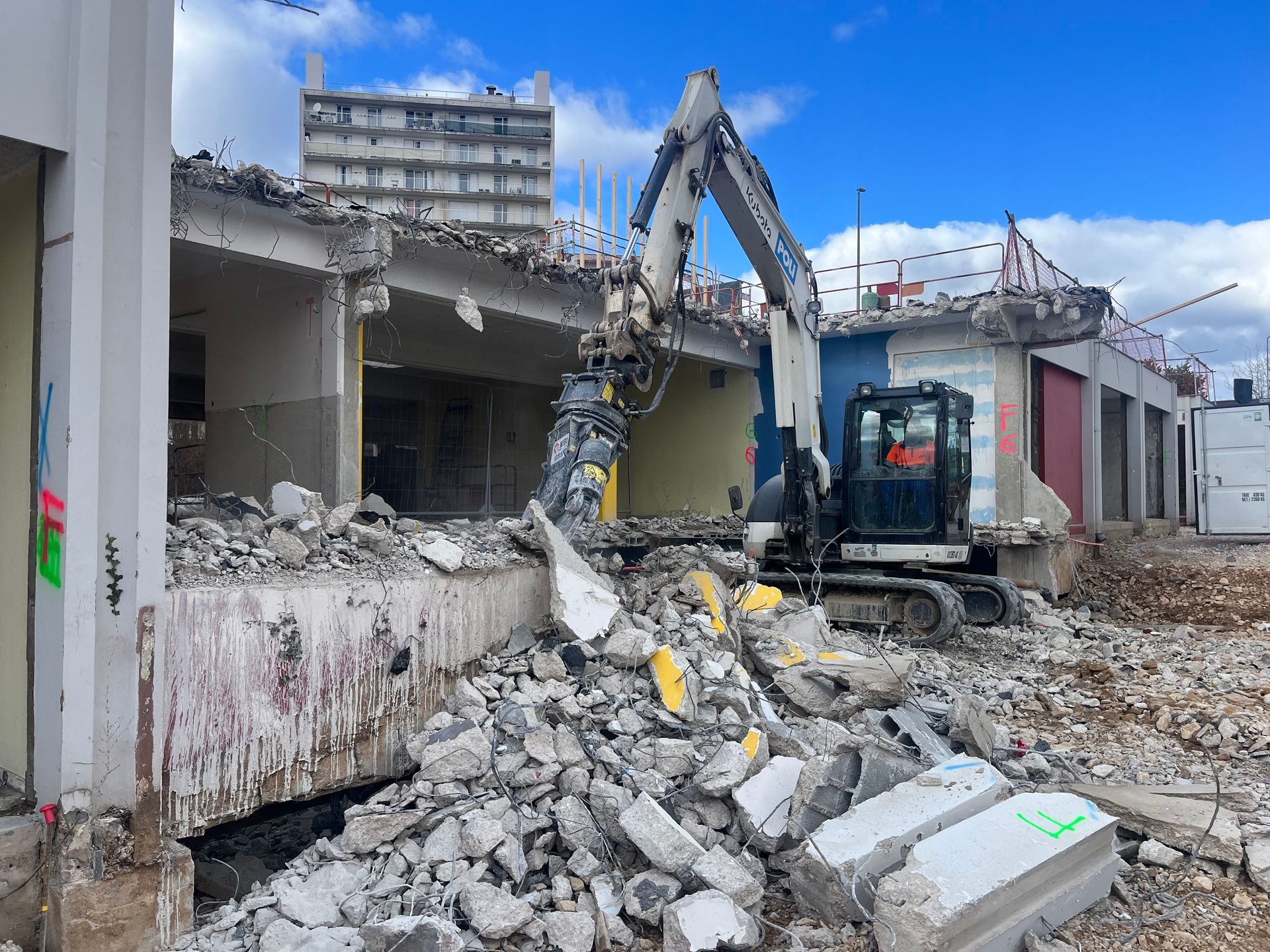 Chantier de démolition avec une pelleteuse en train de travailler sur des débris de construction. Des structures en béton et en métal sont visibles parmi les débris.