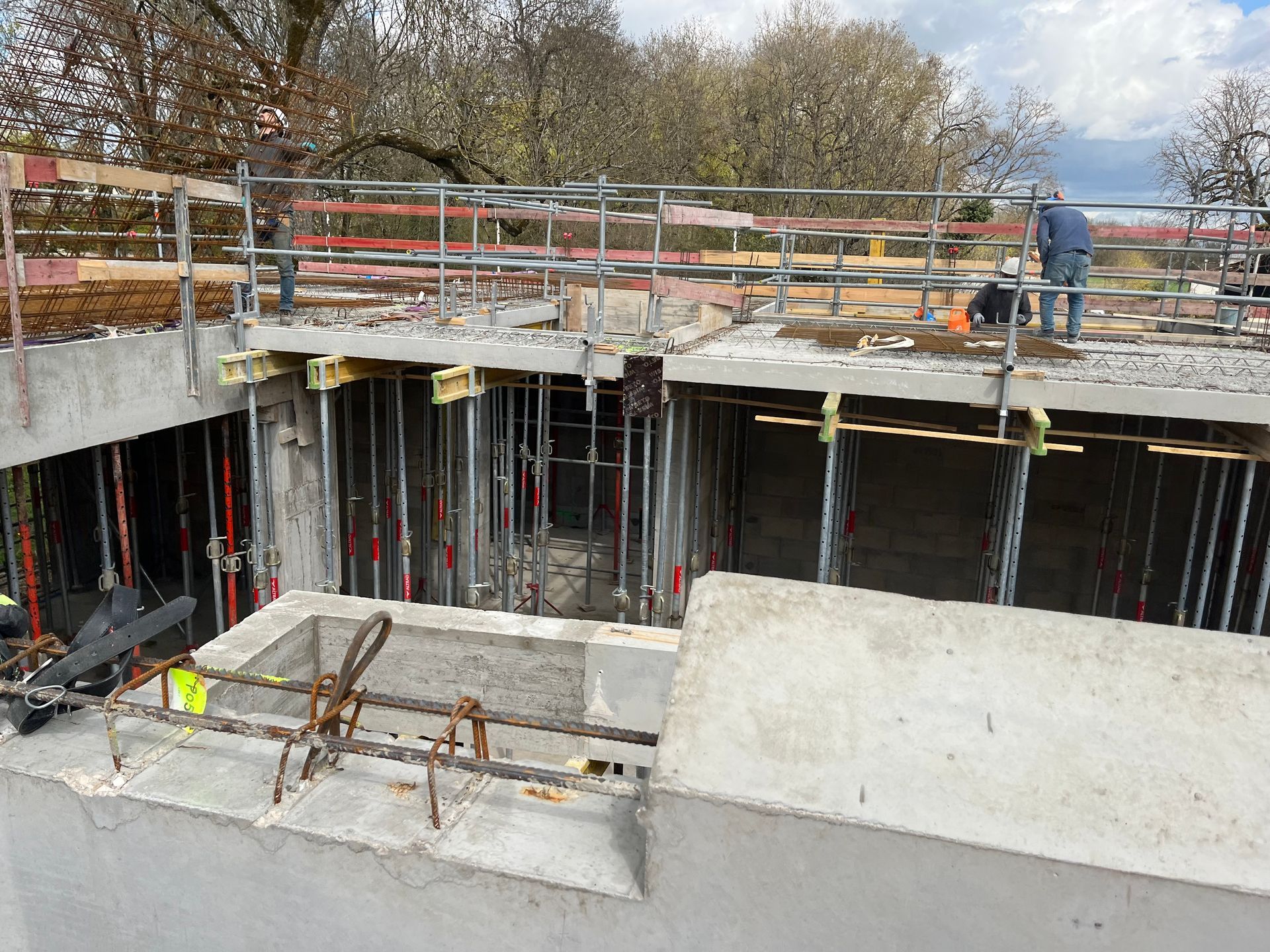 Structure en béton avec des armatures en acier visibles, en cours de construction. Des échafaudages et des barres de renforcement sont visibles.
