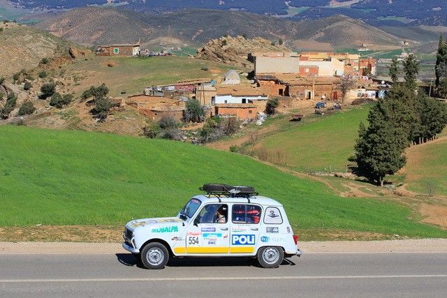Renault 4L sponsorisée roulant sur une route de campagne marocaine avec village en arrière-plan