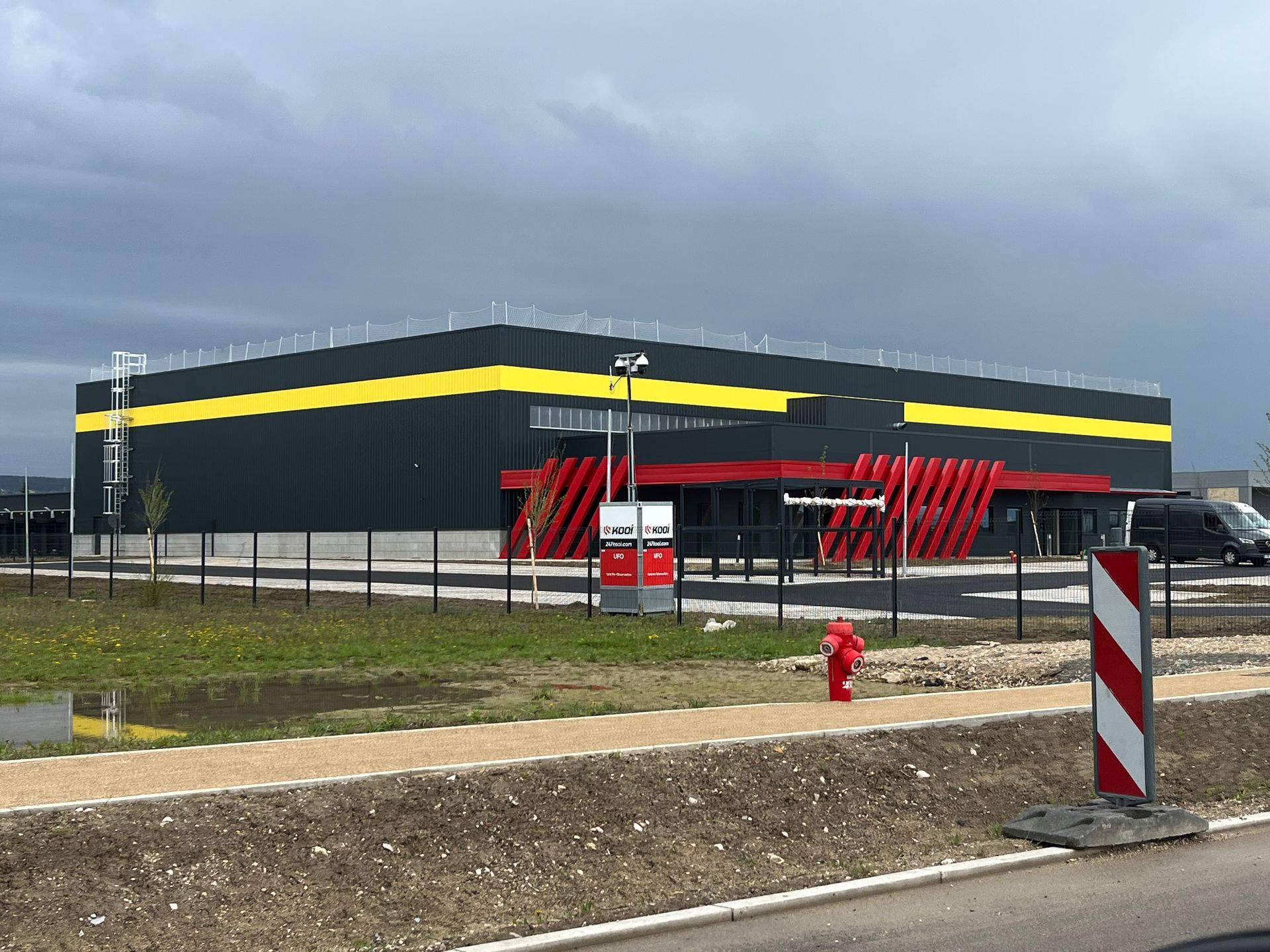 Bâtiment industriel moderne avec des structures en métal et un parking en face. Le ciel est clair et ensoleillé.