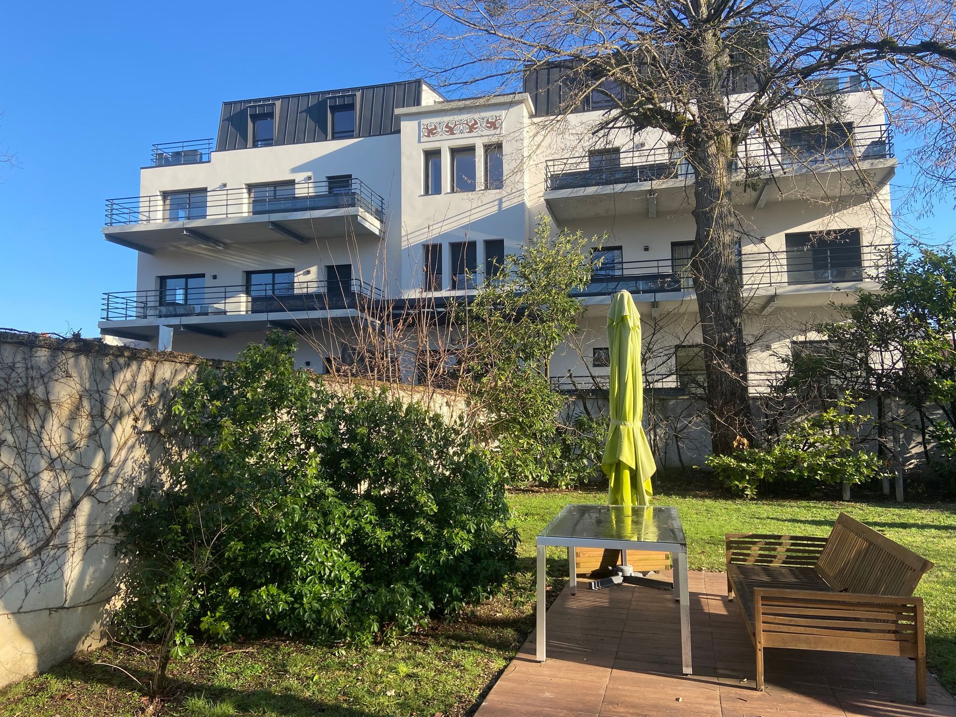 Bâtiment résidentiel moderne avec des balcons et des fenêtres. Le bâtiment est entouré de verdure et le ciel est clair et ensoleillé.