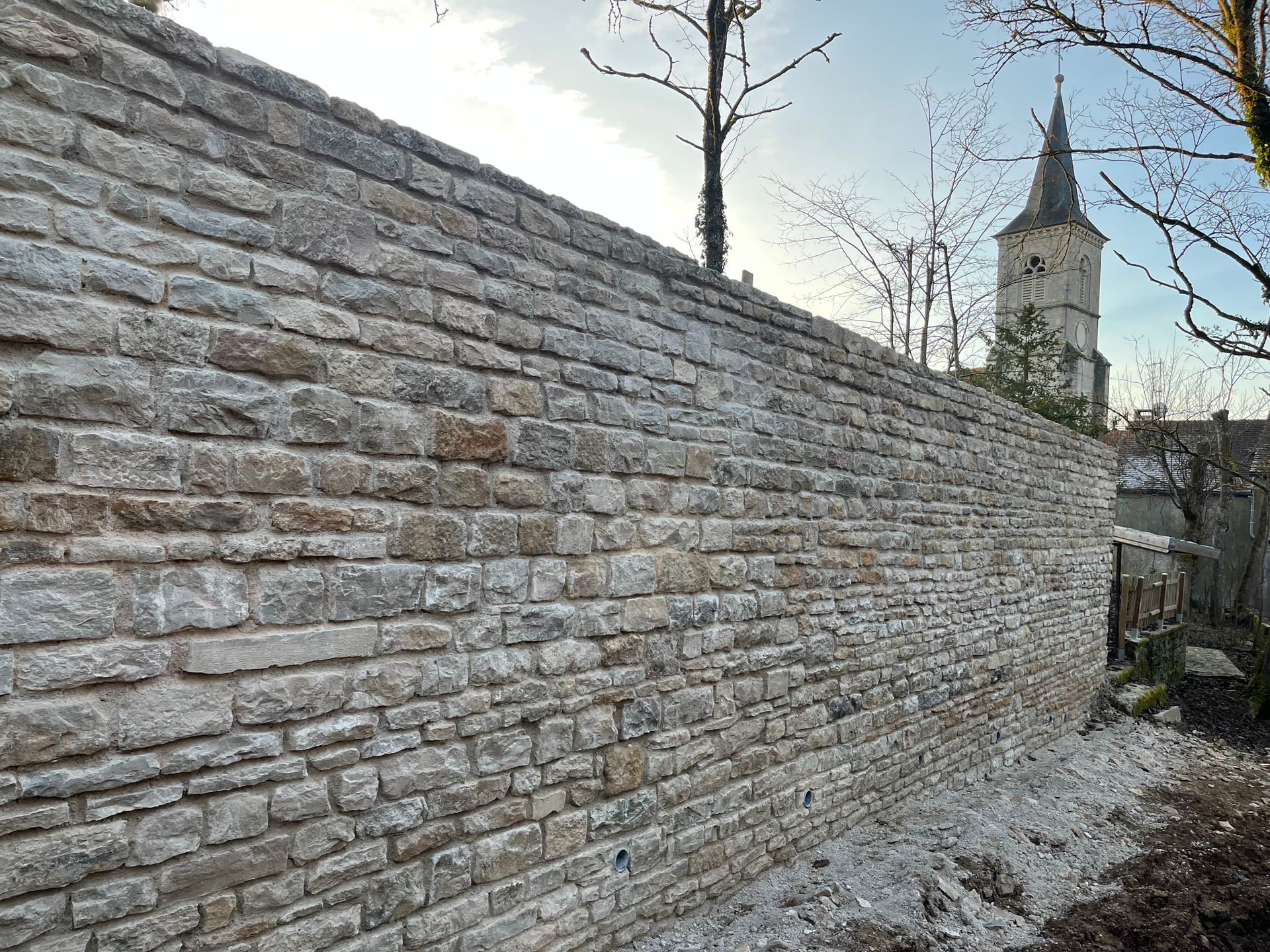 Mur en pierre ancienne avec des détails de construction et un clochet d'Eglise en fond.
