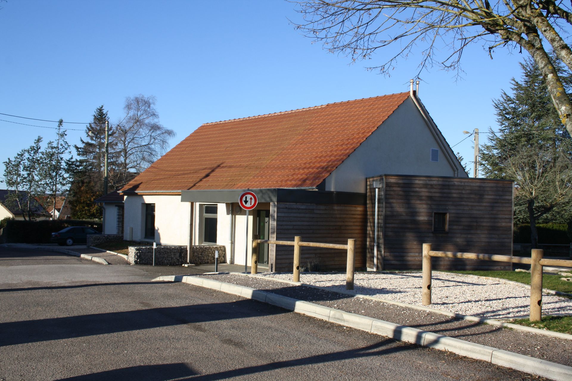 Bâtiment avec un toit en tuiles et un jardin bien entretenu. La maison est entourée de verdure et le ciel est clair et ensoleillé.
