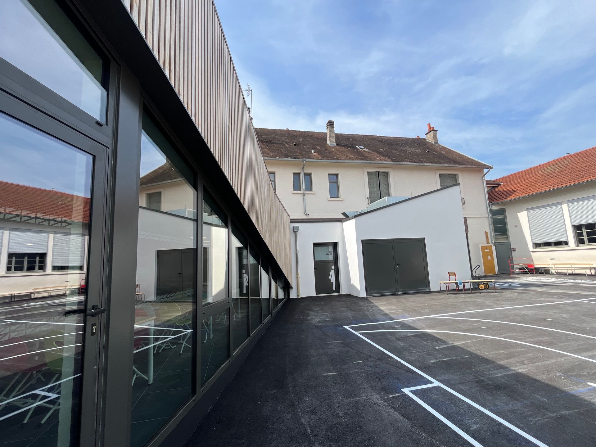 Bâtiment moderne avec une façade en bois et des structures en métal. Le bâtiment est entouré de verdure et le ciel est clair et ensoleillé.