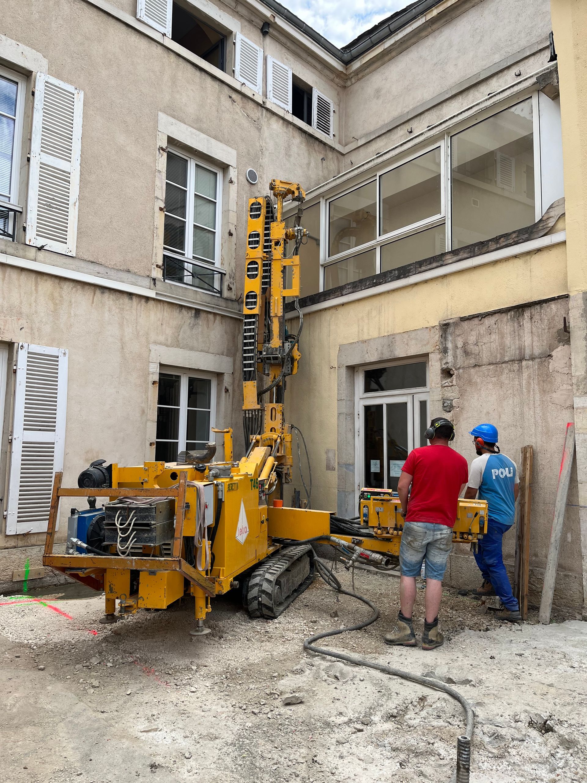 Grue de construction sur un chantier de rénovation. Des ouvriers sont visibles, et le bâtiment en arrière-plan montre des signes de travaux de rénovation.