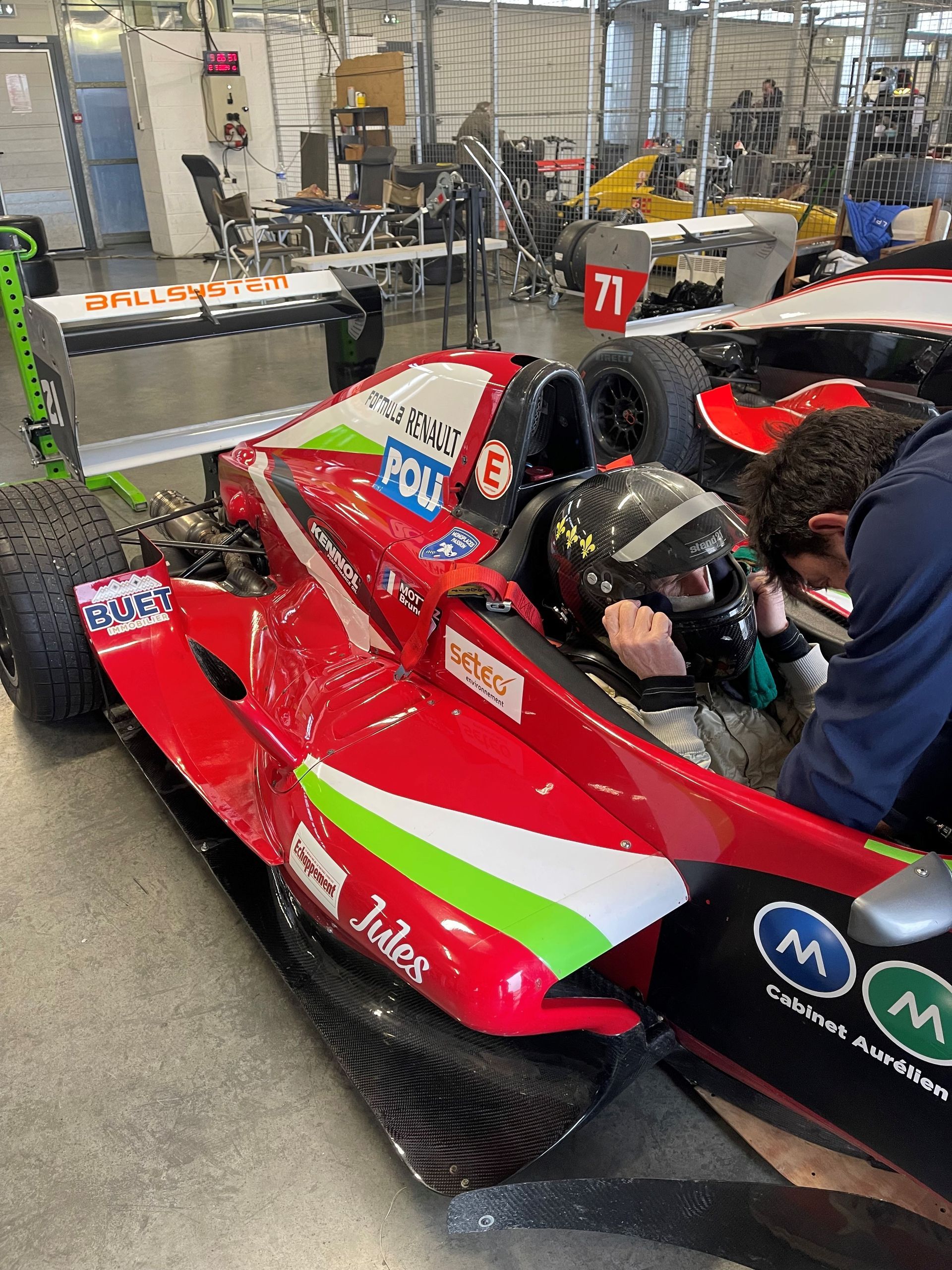 Pilote ajustant son casque dans une monoplace rouge aux couleurs Poli dans un stand avant la course