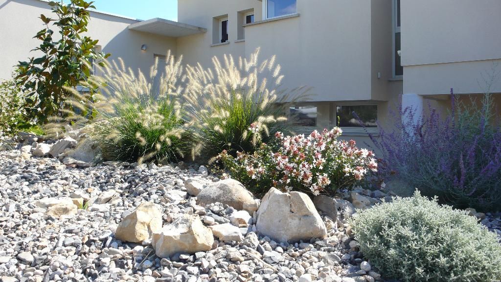 Aménagement paysager avec rochers, herbes et buissons fleuris devant un bâtiment de couleur claire par une journée ensoleillée.
