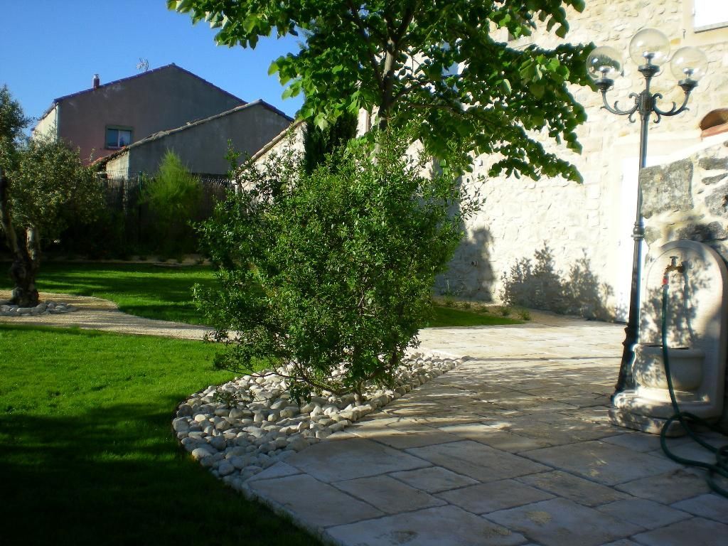 Un espace vert agrémenté d'un buisson et de rochers blancs, flanqué d'un mur en pierre et de pavés, abritant un arbre et deux édifices.