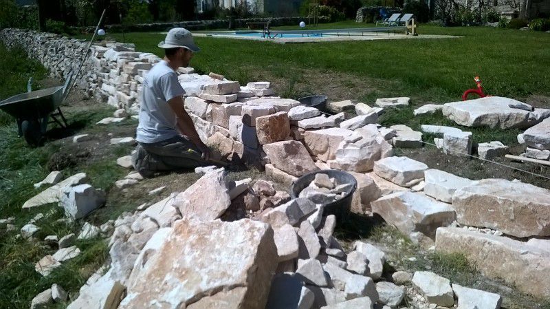 Un homme construit un mur de pierre à l'extérieur près d'une pelouse et d'une piscine, entouré de matériaux de construction.