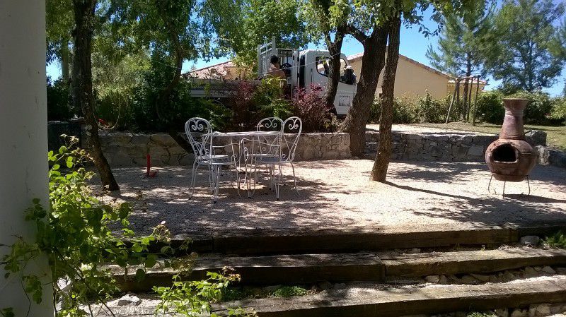 Balcon extérieur équipé d'une table et de chaises en métal blanc, avec des marches en pierre, des arbres et une cheminée.