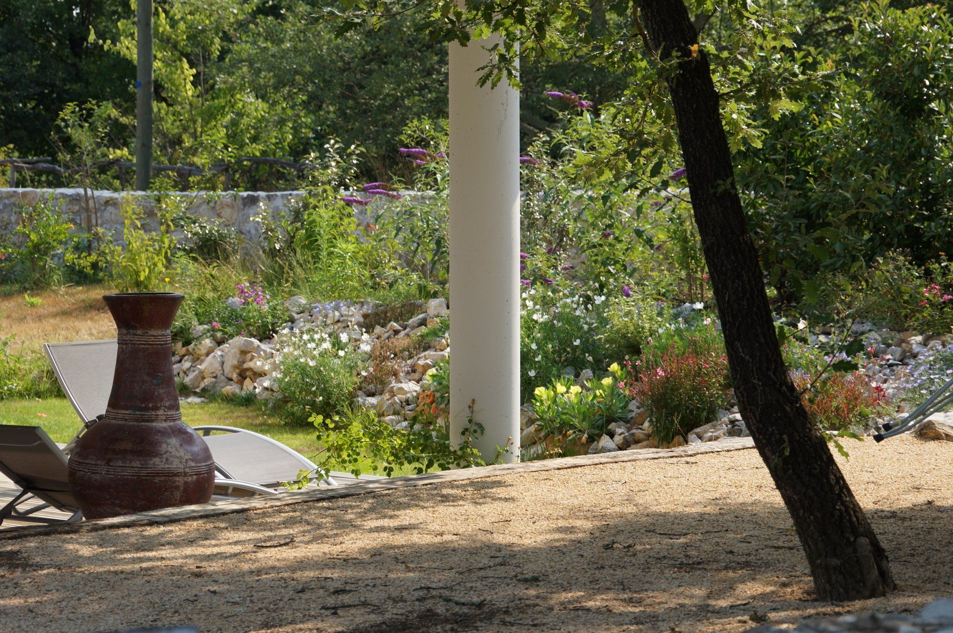 Réalisation de jardin avec un grand vase, un pilier blanc et de la verdure sous l'ombre d'un arbre.