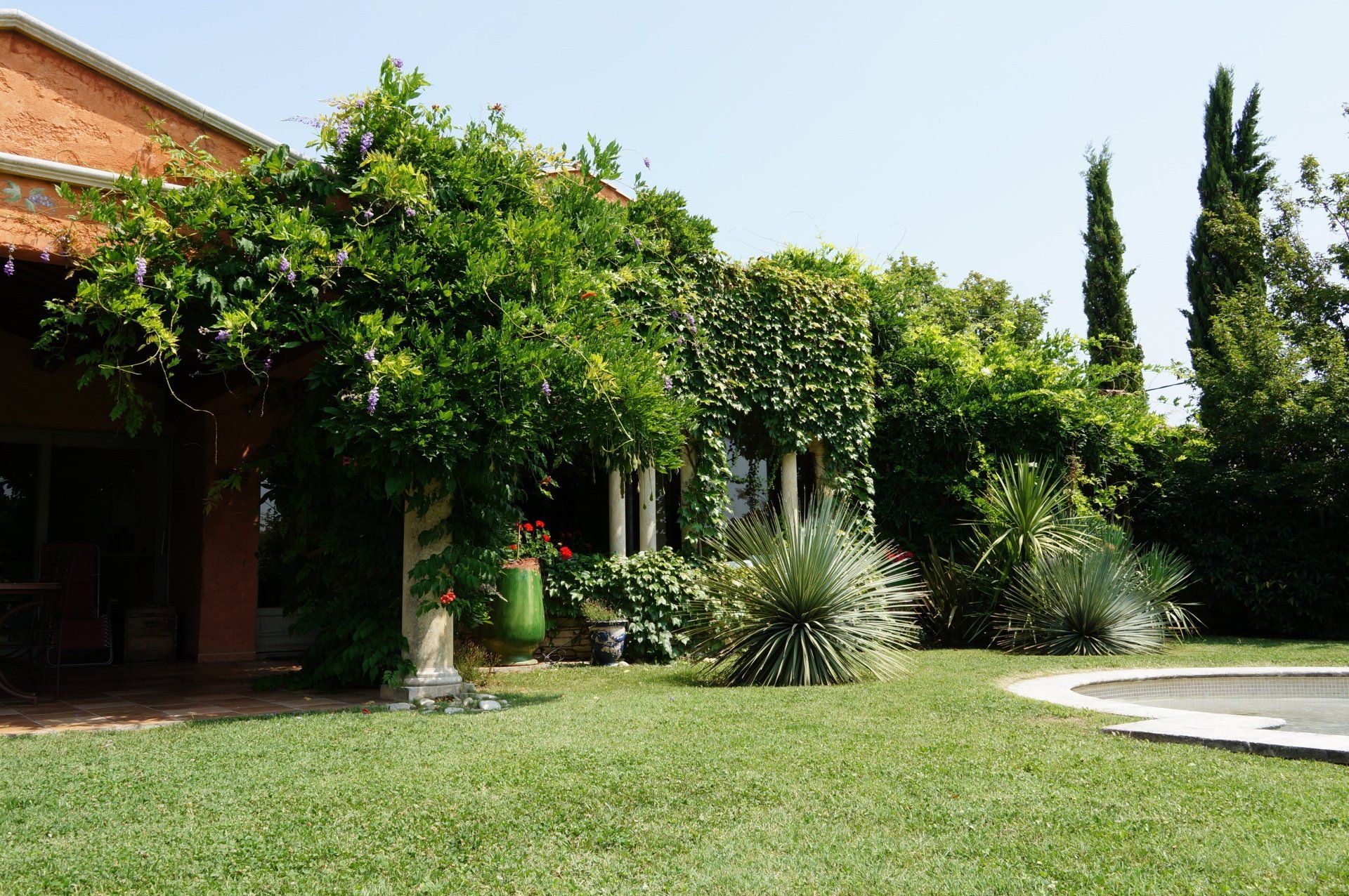 Un jardin verdoyant, orné d'herbe verte et de plantes grimpantes, avec une maison au toit en tuiles argileuses, est illuminé par une journée radieuse.