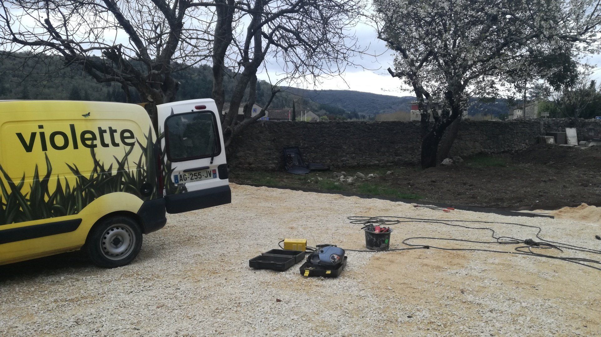 Véhicule utilitaire de l'entreprise de couleur jaune garé sur le gravier du chemin entouré d'un jardin, avec des outils à l'extérieur.