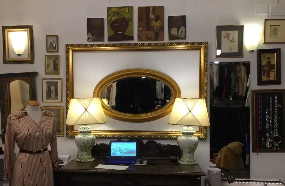Interior de una tienda vintage con un espejo ornamentado, arte enmarcado y una exhibición de ropa y joyas.