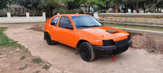 Automóvil hatchback modificado de color naranja y negro estacionado en un camino de tierra al lado de una pared de ladrillos.