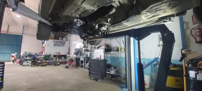 Un coche se eleva en un elevador dentro de un taller mecánico. Se ve el chasis y el taller está desordenado.