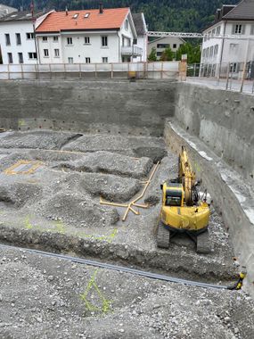 Zwei Bauarbeiter in orangefarbenen Westen stehen auf einer Erdbaustelle neben einer Spritzbeton-Stützmauer.