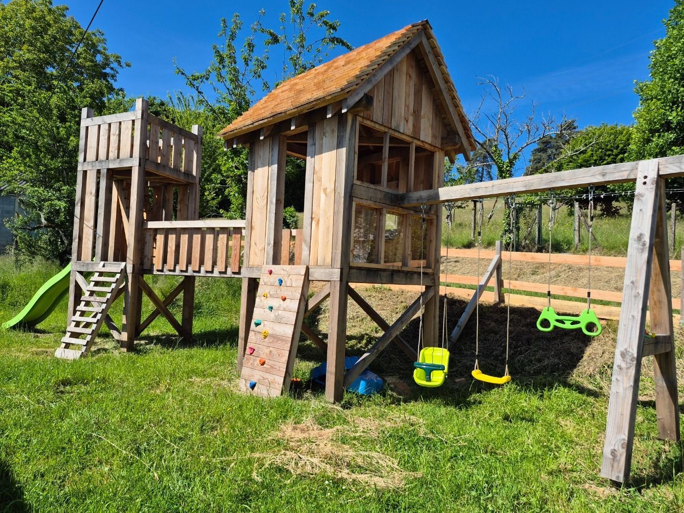 Cabane de jardin en bois avec toboggan, échelle, balançoires et mur d'escalade par une journée ensoleillée.