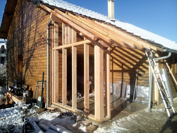 Extension de maison en bois partiellement construite, avec charpente apparente et neige au sol.