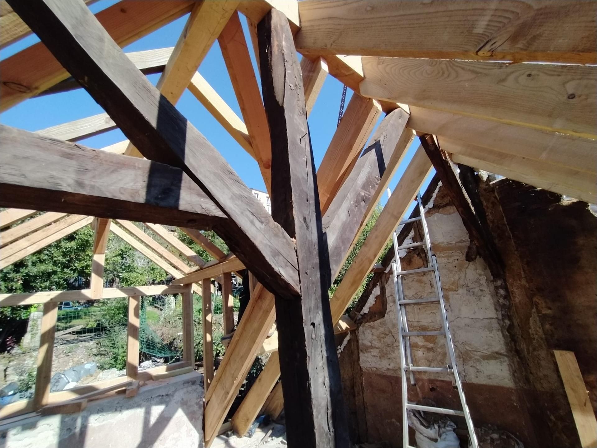 Charpente de toit en bois en construction, avec une échelle et le ciel bleu visible à travers les poutres.