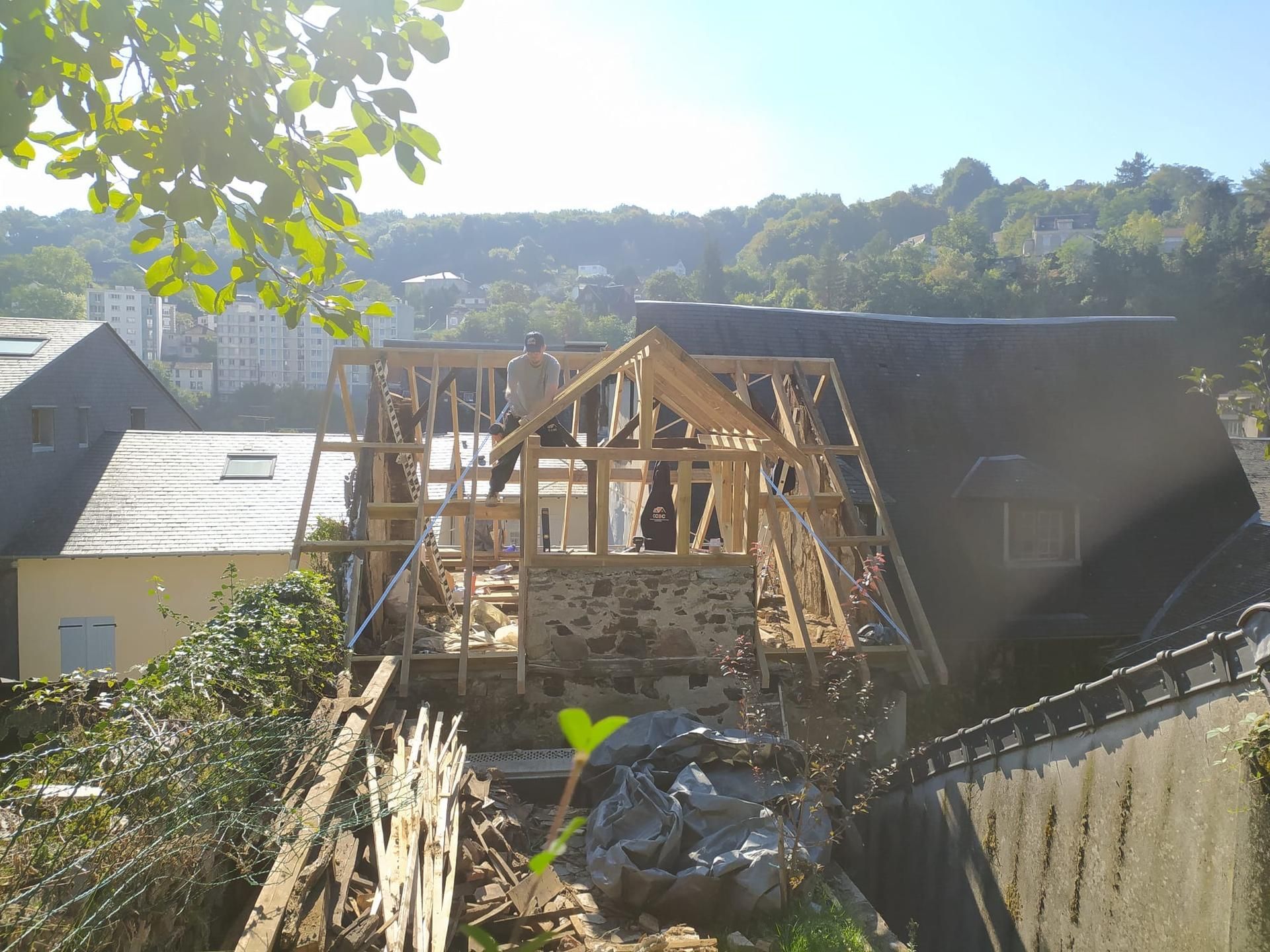Maison à ossature en bois en construction à côté d'un mur de pierre, avec des gravats et des arbres baignés de soleil.