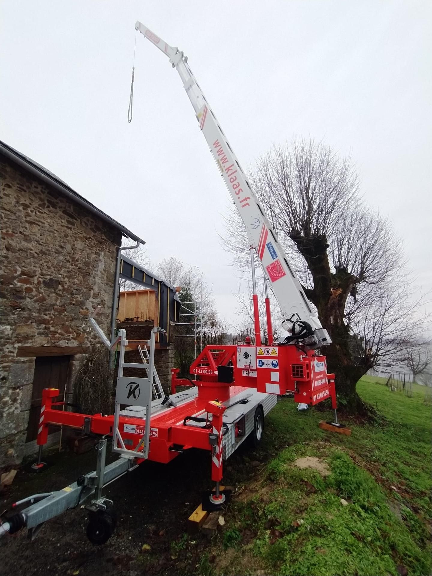 Un gros camion-grue rouge soulève une longue flèche blanche à côté d'un bâtiment en pierre dans une cour herbeuse.