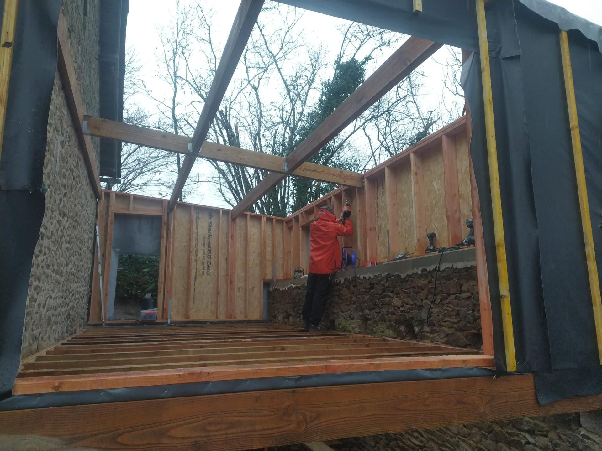 Une personne se tient à l'intérieur d'une structure en bois partiellement construite, avec des poutres.