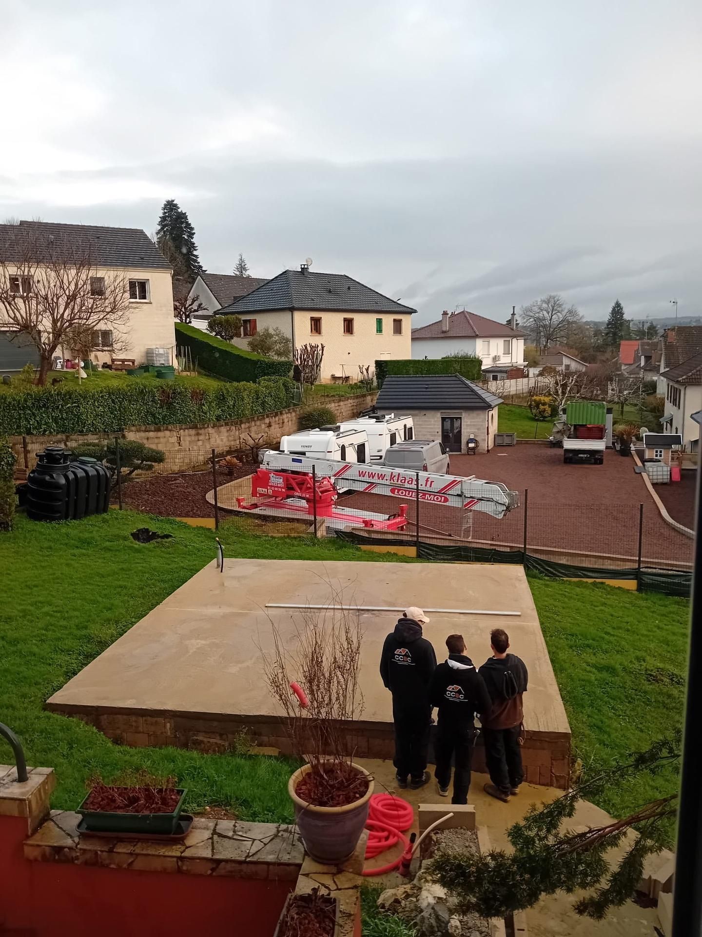 Trois personnes se tiennent debout dans un jardin, à l'arrière d'une maison, sous un ciel gris.