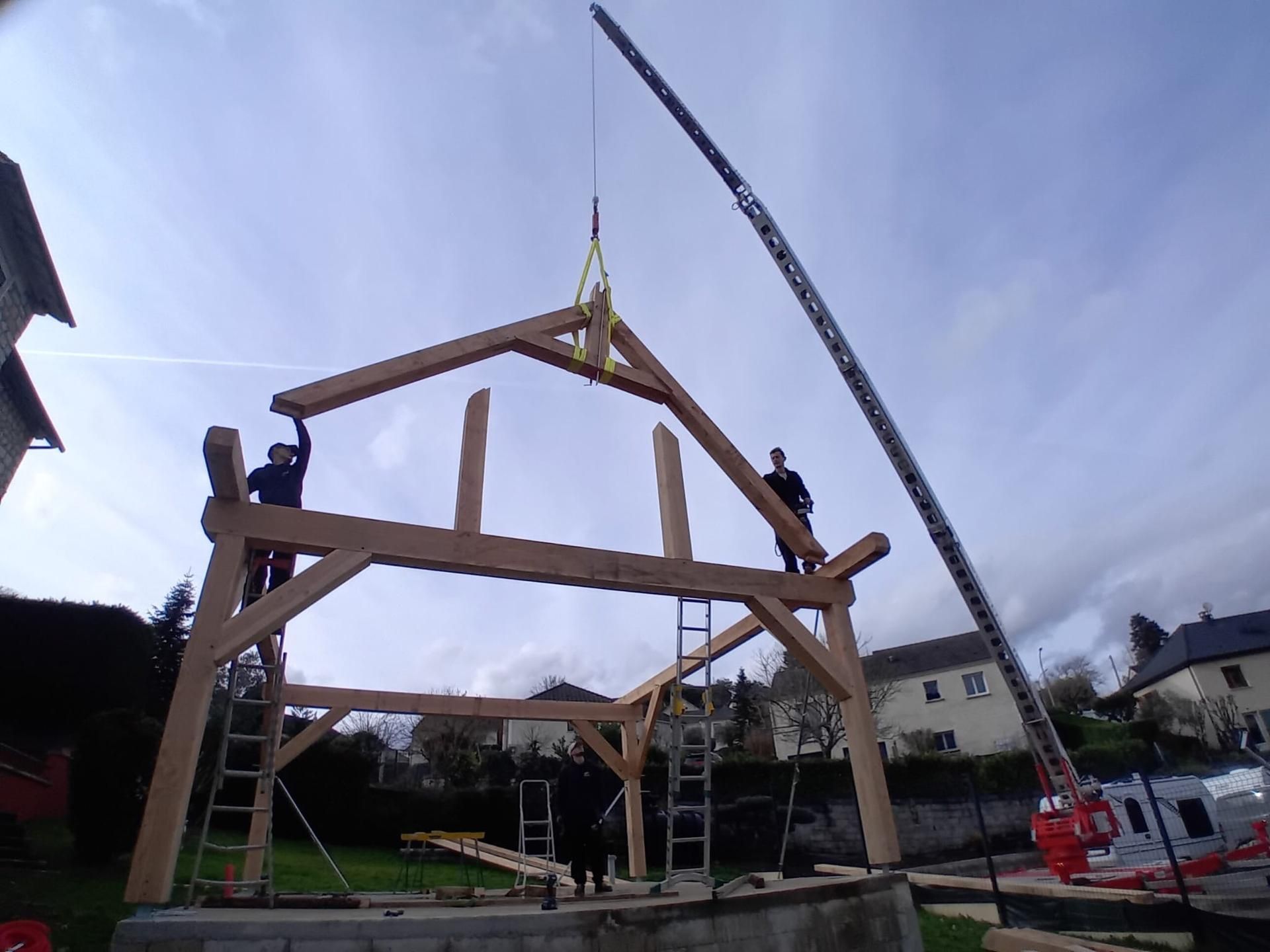 Des ouvriers hissent une grande charpente en bois sur un chantier de construction à l'aide d'une grue.