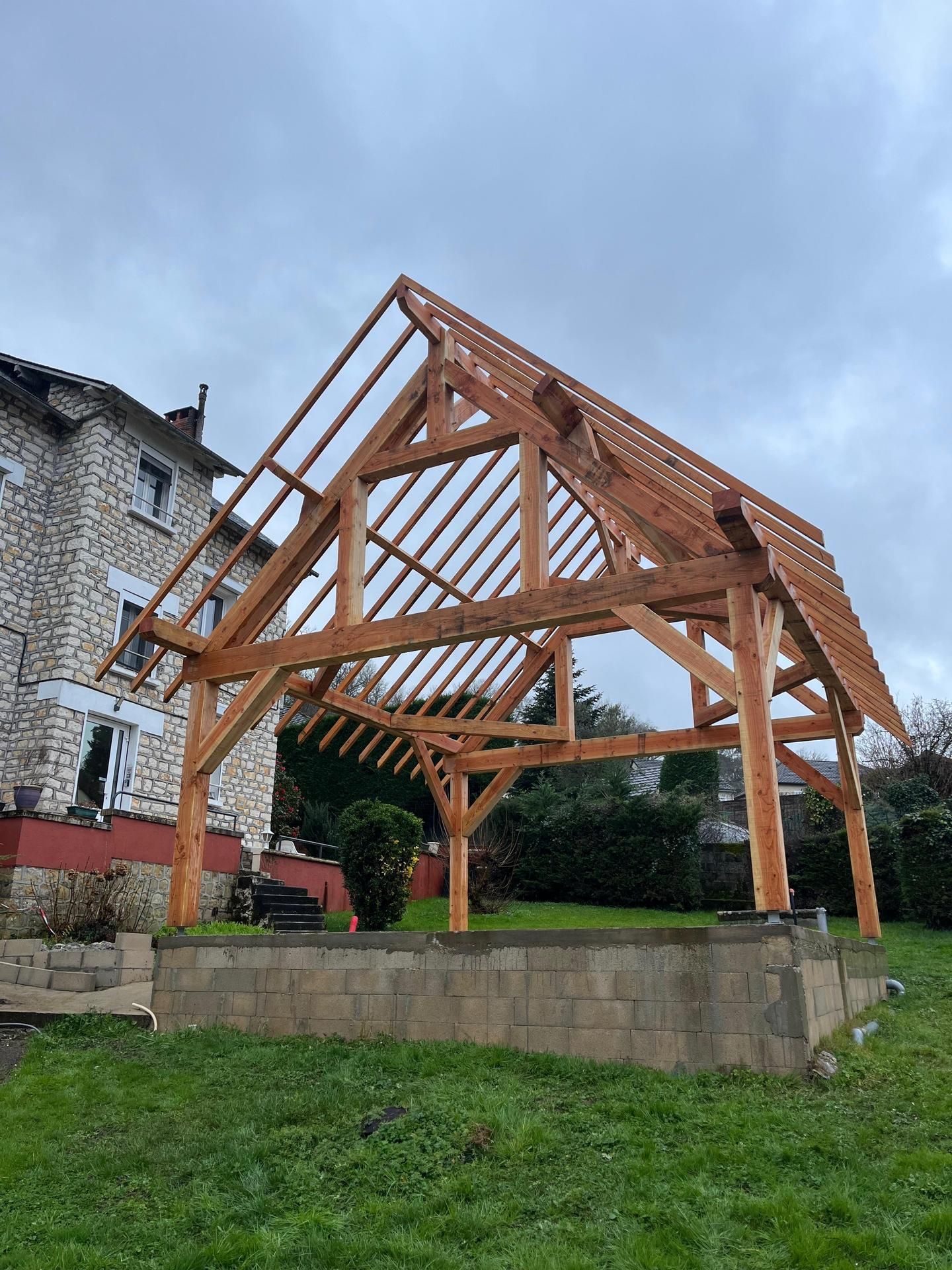 Pavillon à ossature bois en construction à côté d'un bâtiment en pierre et d'une haie, sur une pente herbeuse.
