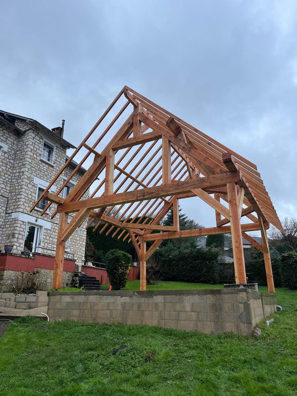 Pavillon à ossature bois en construction à côté d'un bâtiment en pierre et d'une haie, sur une pente herbeuse.