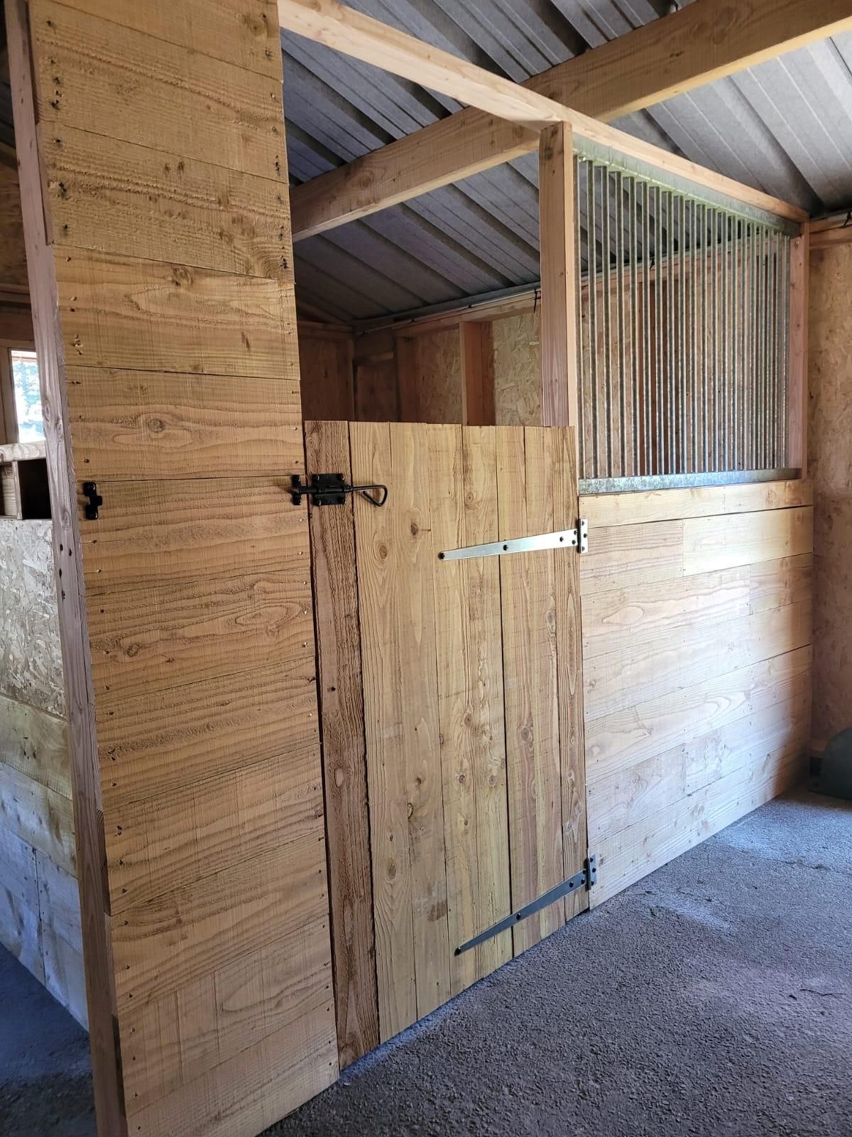 Box d'écurie en bois avec portes en contreplaqué, panneaux muraux ondulés et sol en gravier.