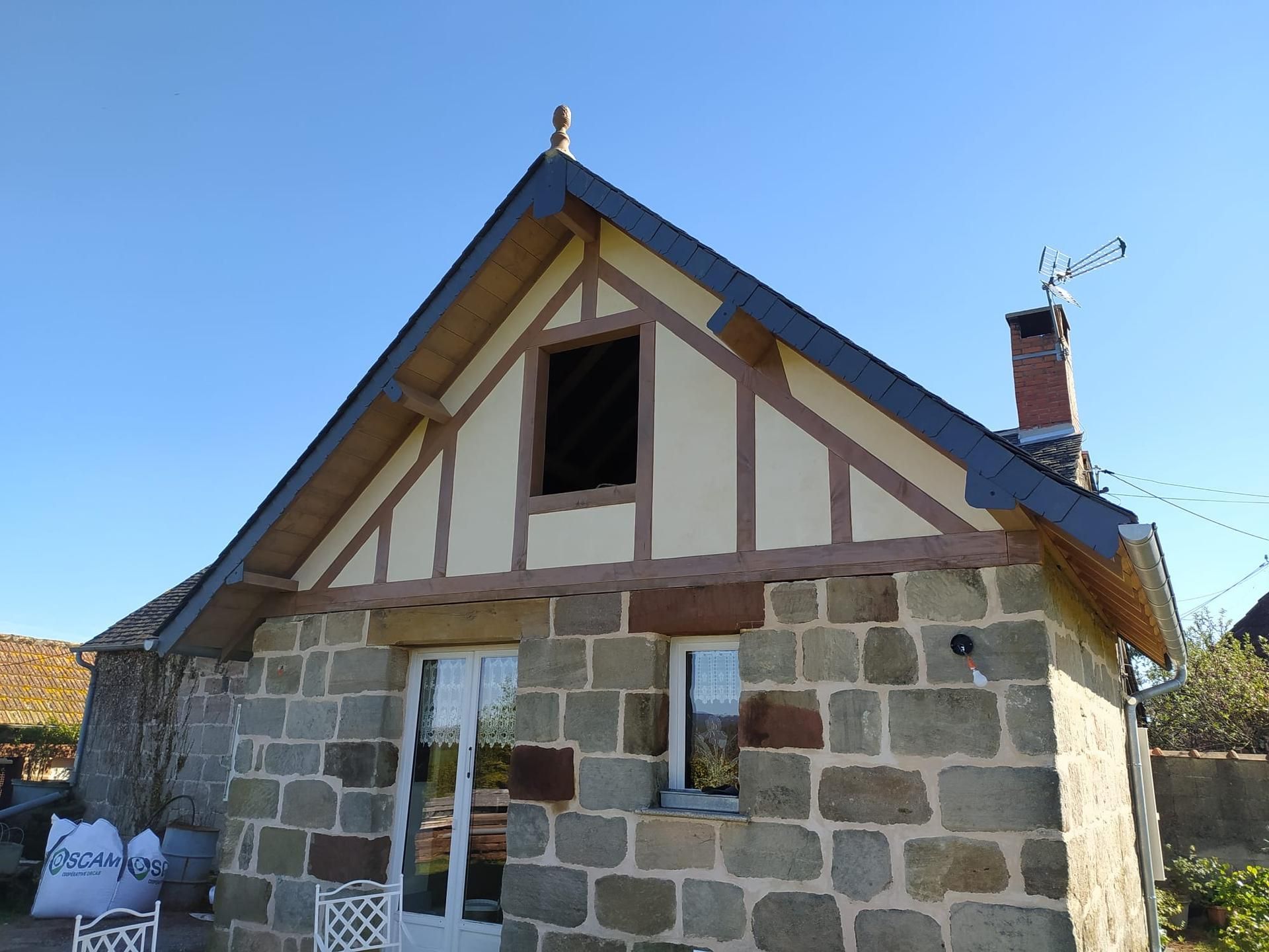 Maisonnette en pierre au toit bleu pentu, aux petites fenêtres et à la cheminée en briques, sous un ciel dégagé.