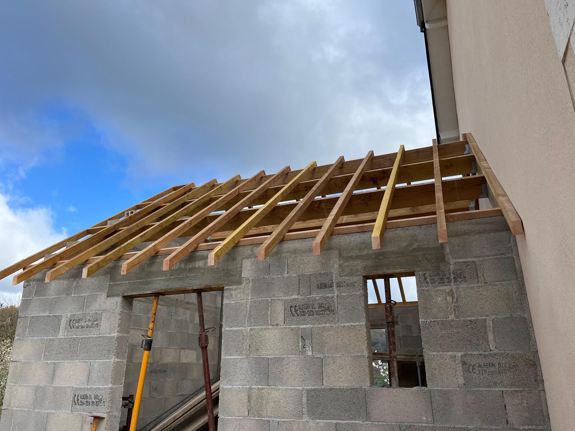 Maison en parpaings partiellement construite, avec charpente apparente, sous un ciel nuageux.