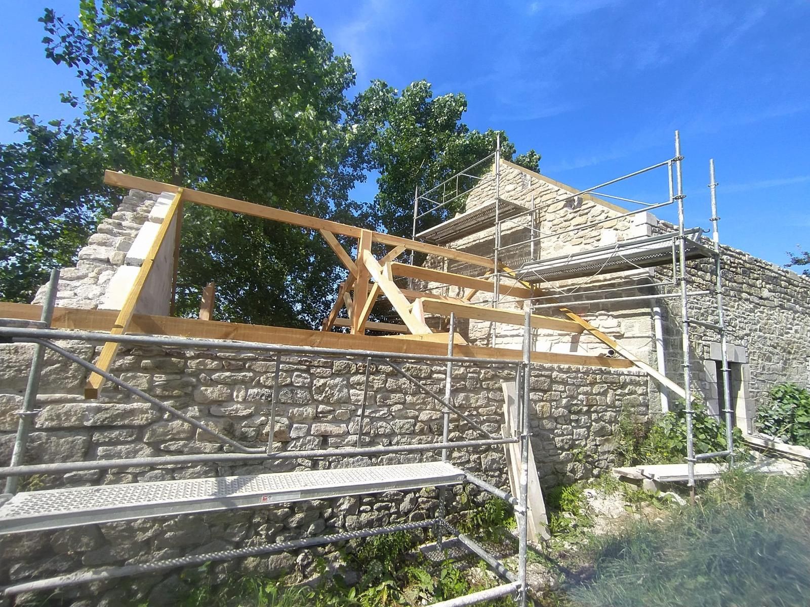 Bâtiment en pierre en rénovation, avec des échafaudages en bois et un mur de pierre incliné sous un ciel bleu.