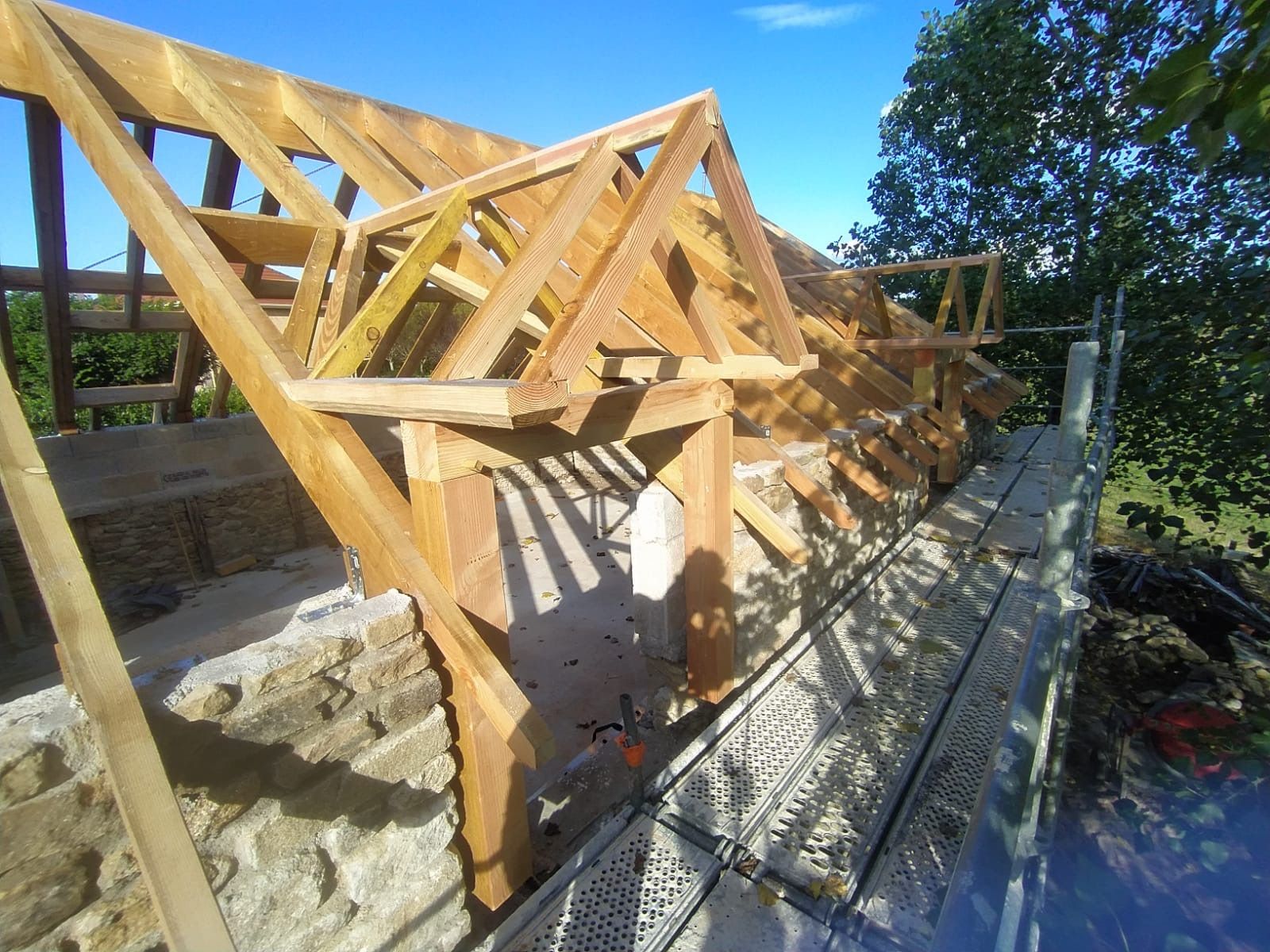 Charpente de toit en bois en construction sur un mur de pierre, avec ciel bleu et arbres en arrière-plan.