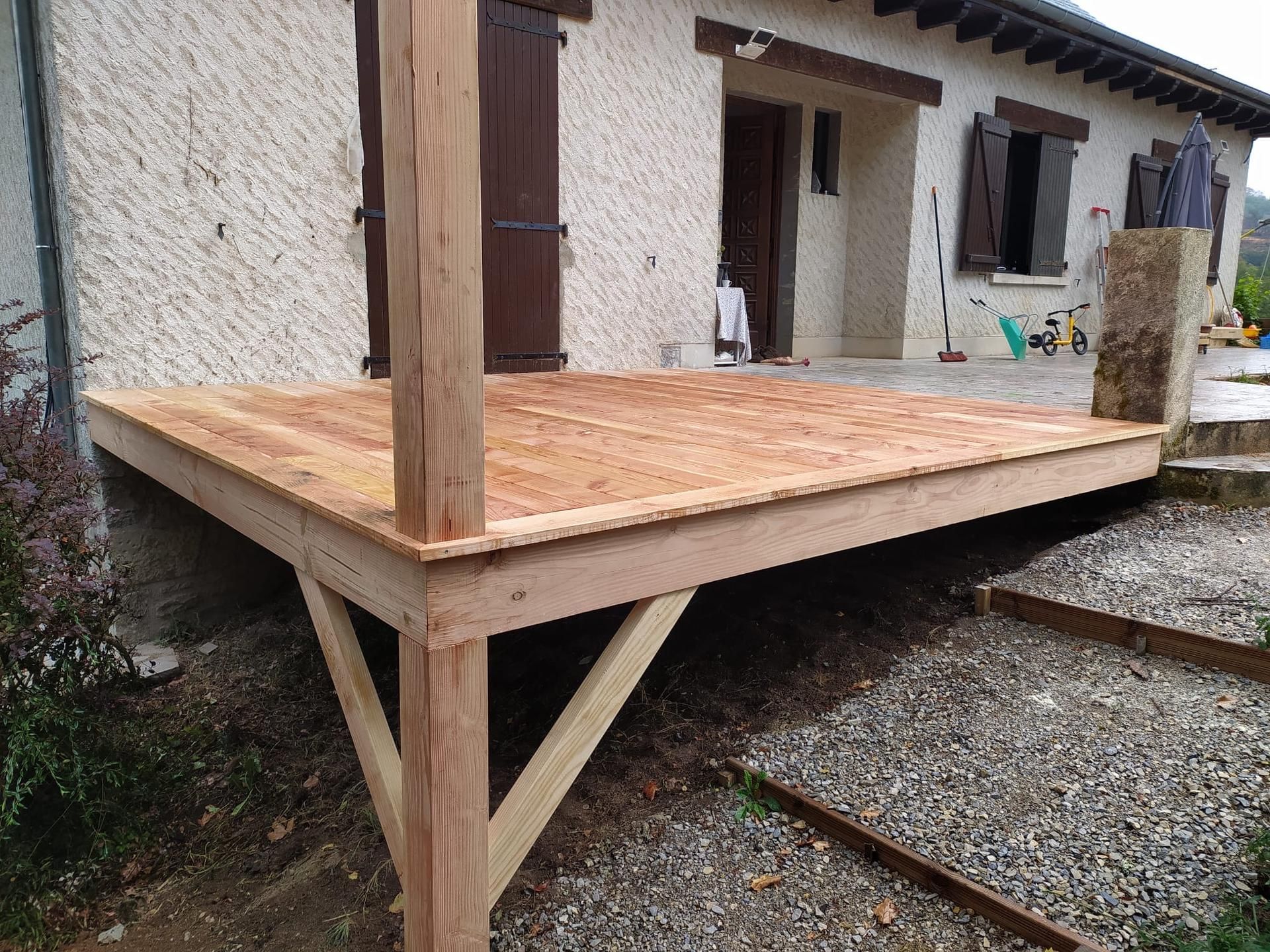 Terrasse en bois avec poteaux à côté d'une maison en stuc et d'une cour en gravier.