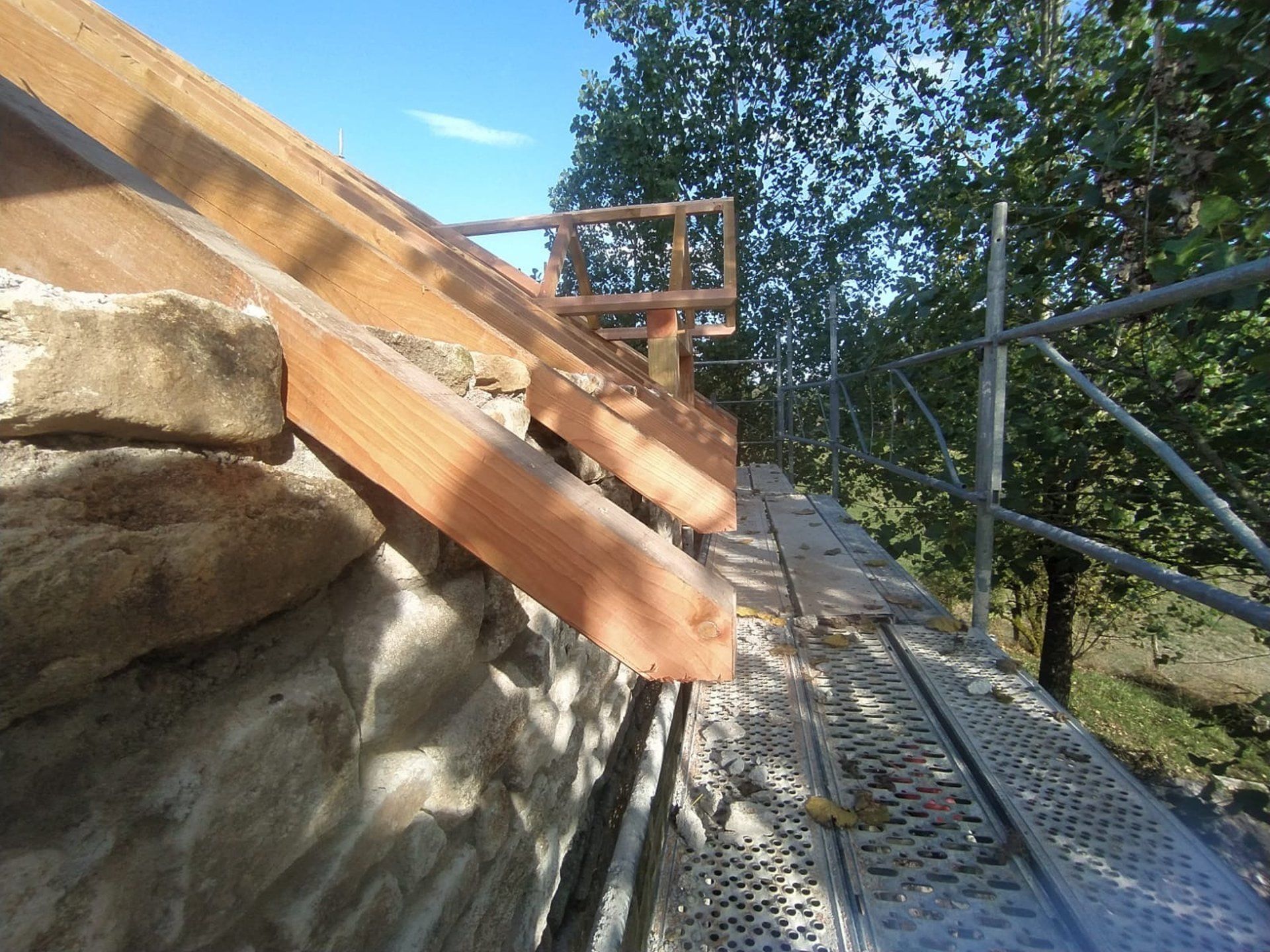 Échafaudage à côté d'un escalier en bois adossé à un mur de roche et à des arbres, en extérieur.
