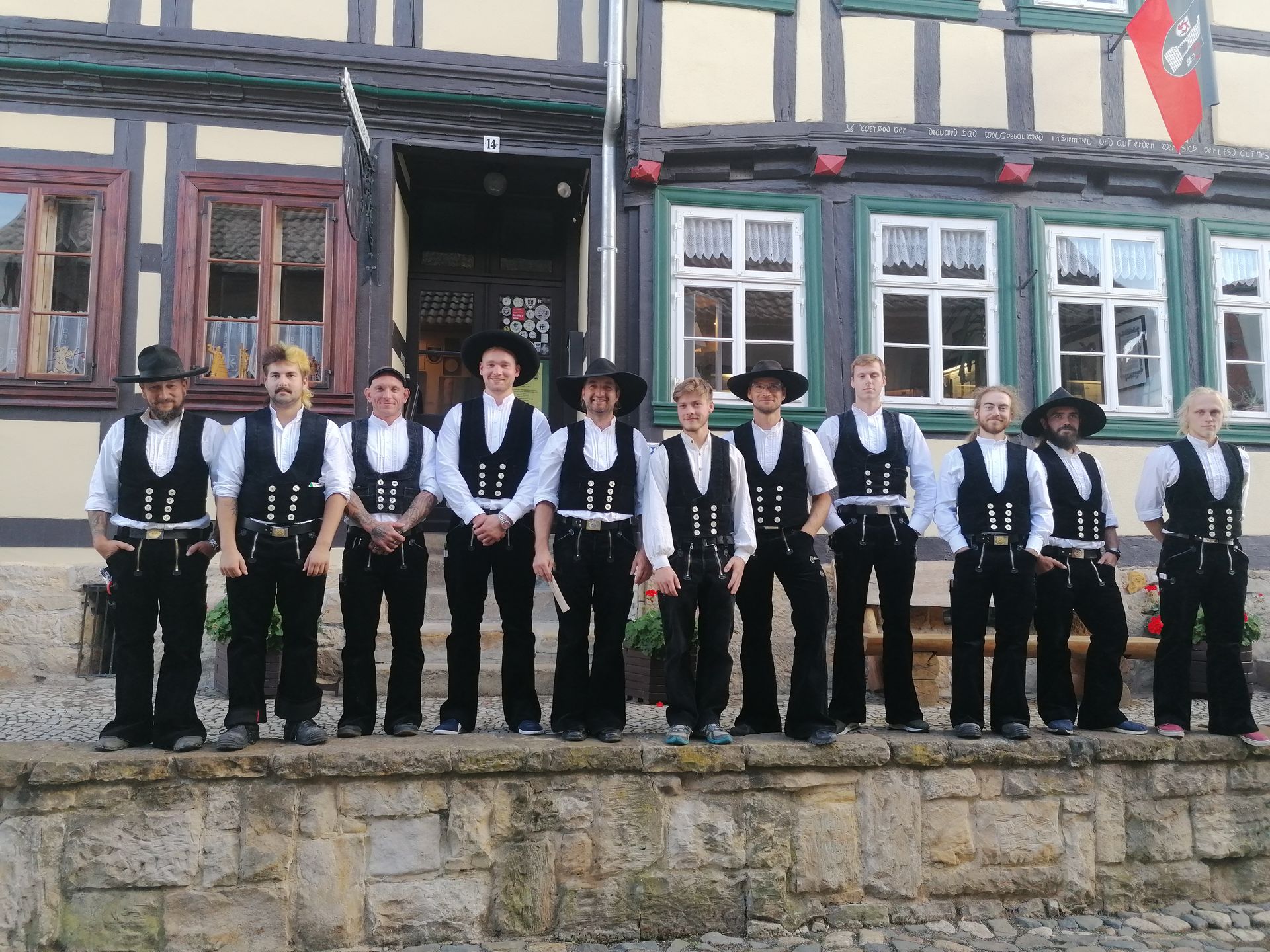 Gruppe von elf jungen Zimmermännern auf der Walz in Zunftkleidung auf einer Mauer vor einem Holzhaus