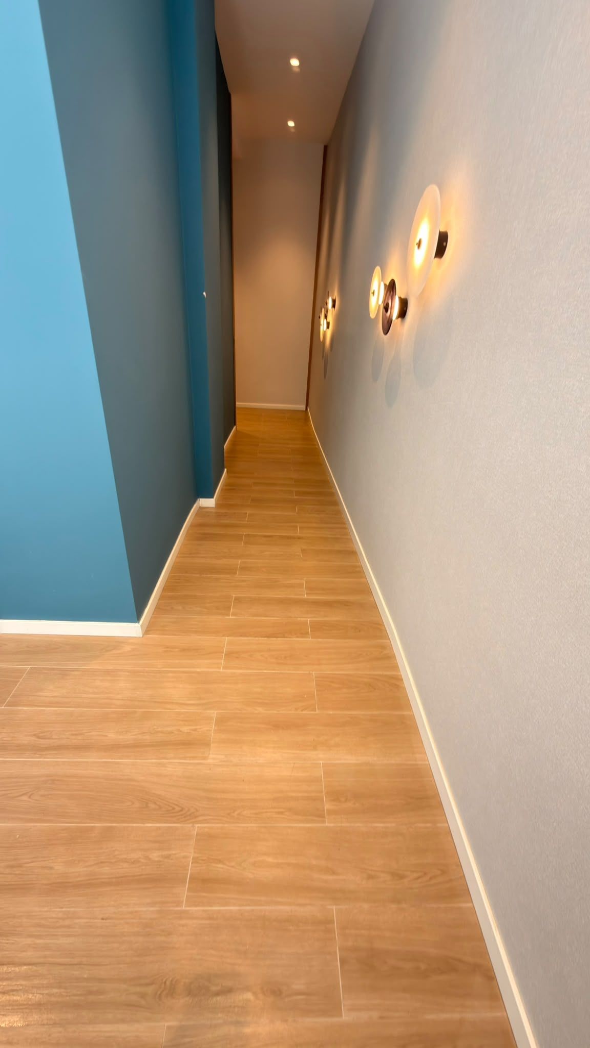 Pasillo interior estrecho con suelo de madera, paredes blancas, una pared azul de acento y apliques de pared.