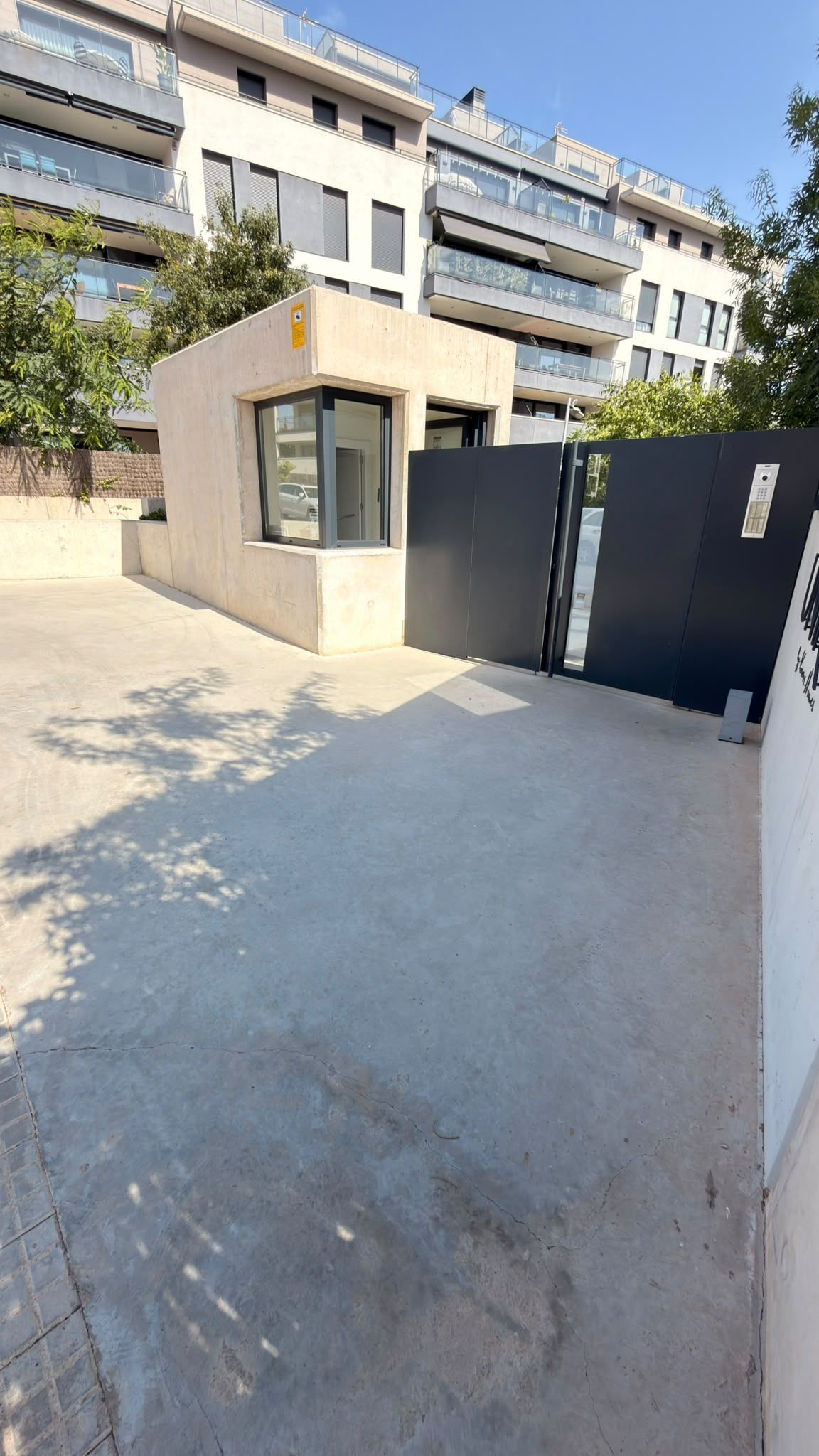 Entrada con portón a un moderno complejo de apartamentos, con una pequeña garita de vigilancia y una entrada pavimentada