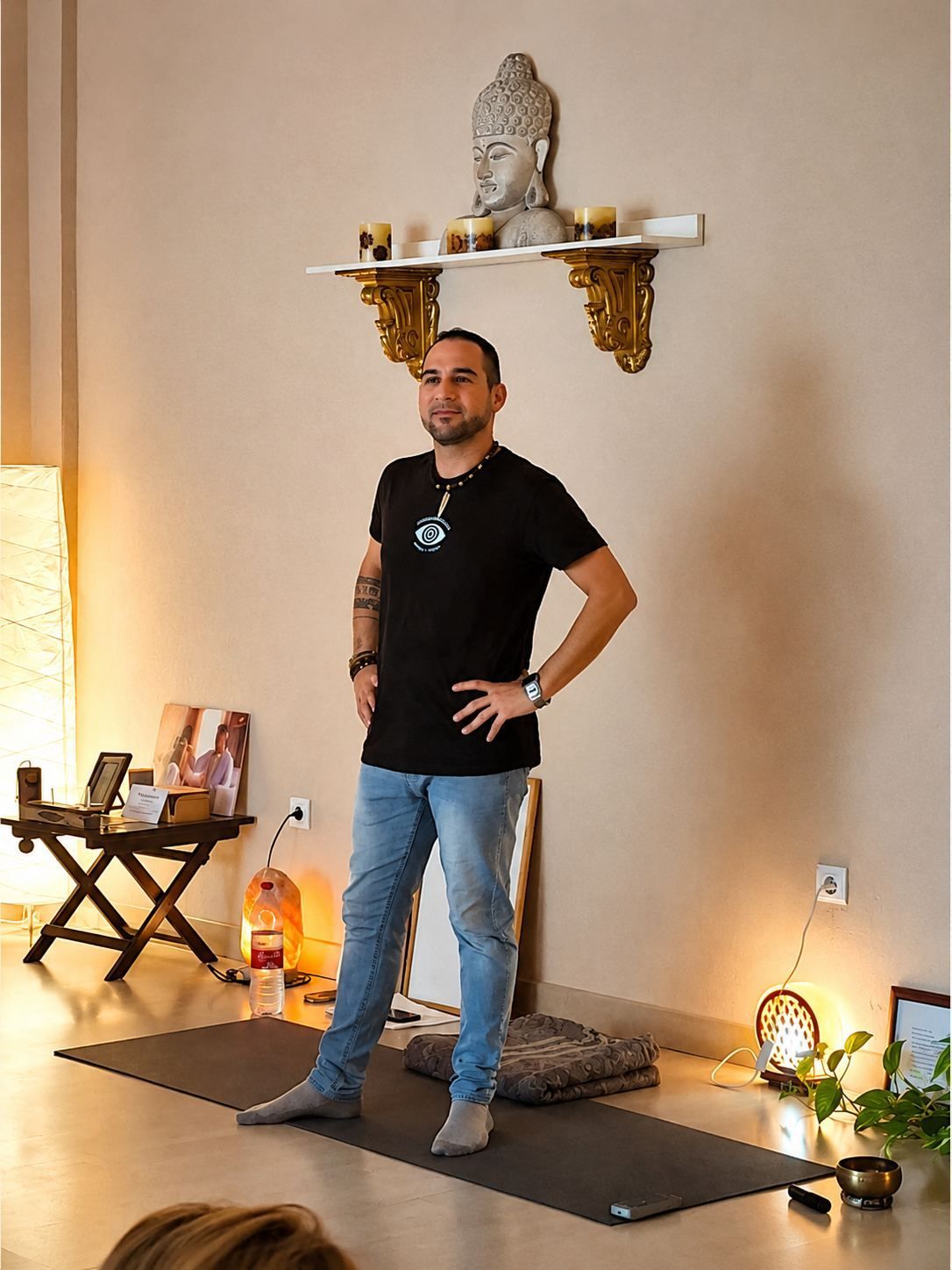Hombre posando en el interior sobre una esterilla de yoga, vistiendo una camisa negra y vaqueros.